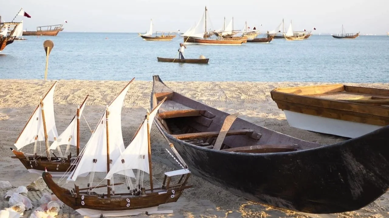 Traditional wooden dhow boats and miniature dhow models with white sails resting on a sandy beach in Qatar, with active seafaring boats on the calm blue water in the background.