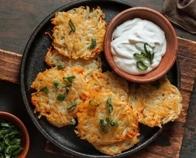 Golden-brown potato latkes served on a black plate with a side of sour cream and chopped green onions.