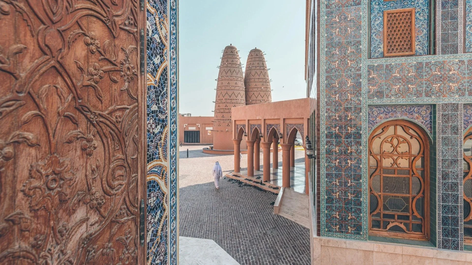 A view through an intricately carved wooden door frame showing the blue-tiled walls of Katara Mosque and its distinct tall pigeon towers in the distance.