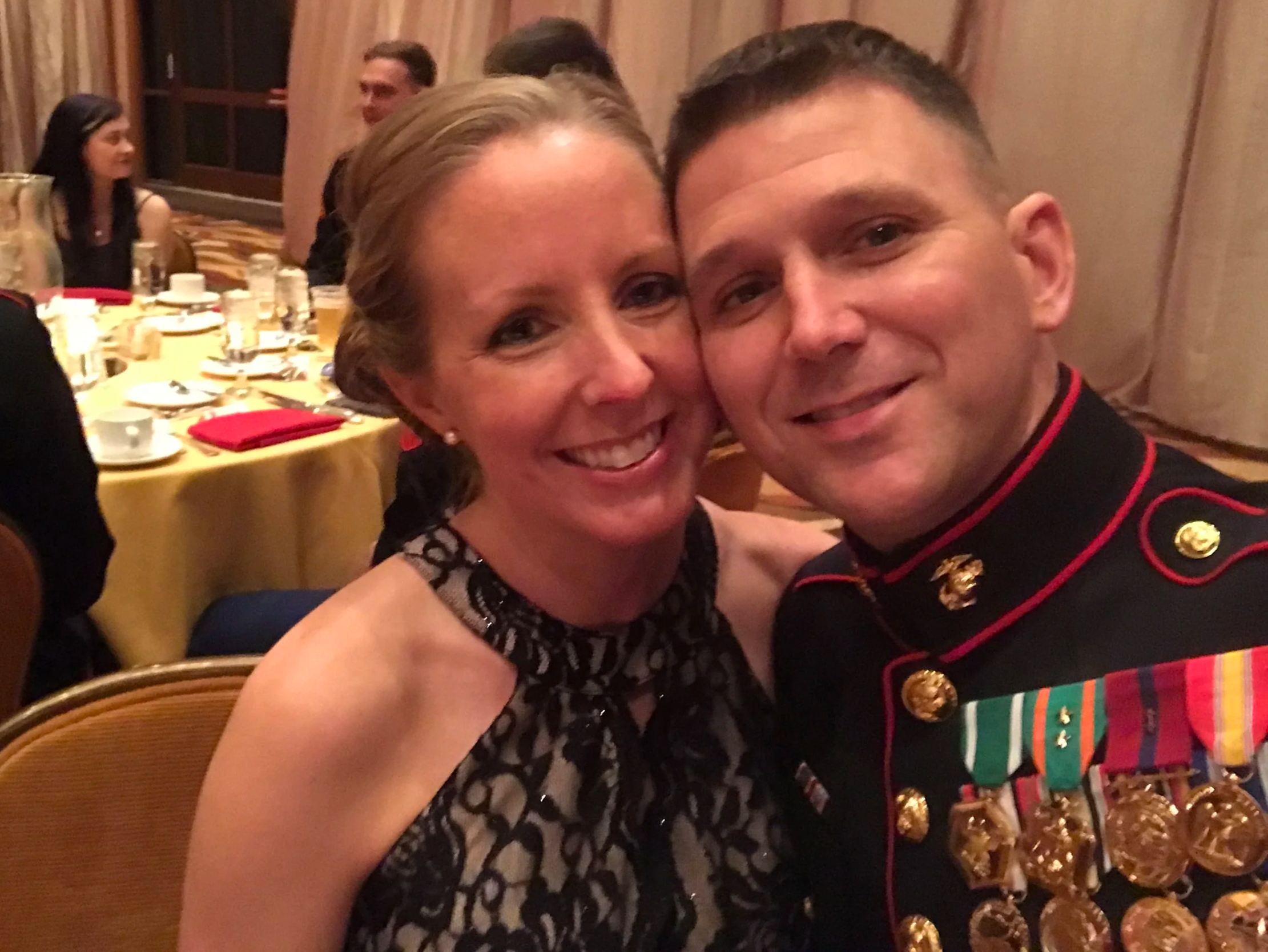 A woman and a man taking a close-up selfie at a formal event or dinner, with the woman wearing a black patterned dress and the man in a decorated military uniform with medals.