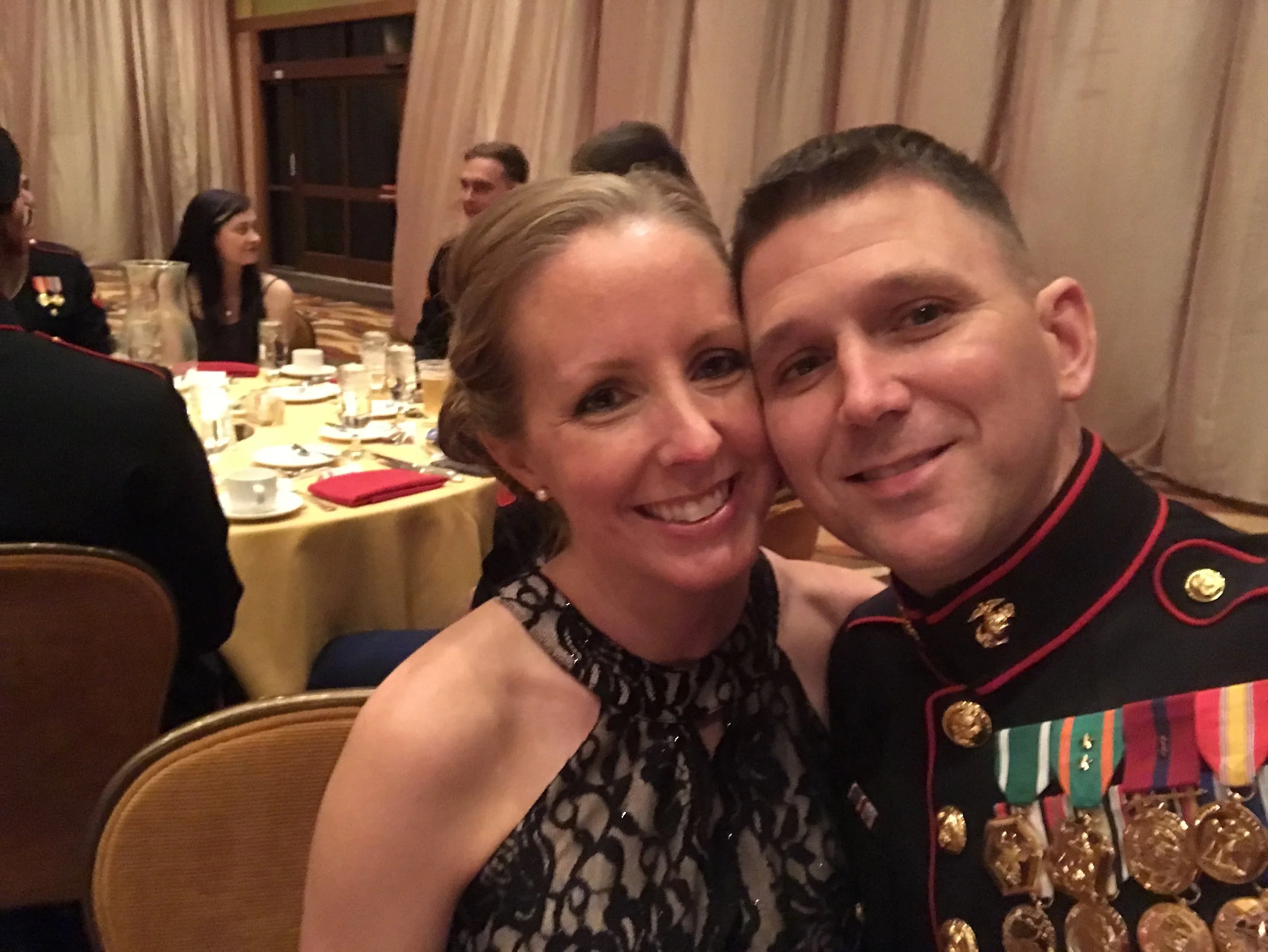 A woman in a black lace dress and a man in a military uniform with medals are smiling and taking a selfie at a formal dinner event.
