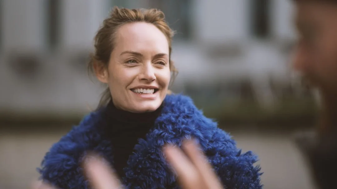 A woman smiling and talking to a person who is partially visible in the foreground, outdoors.