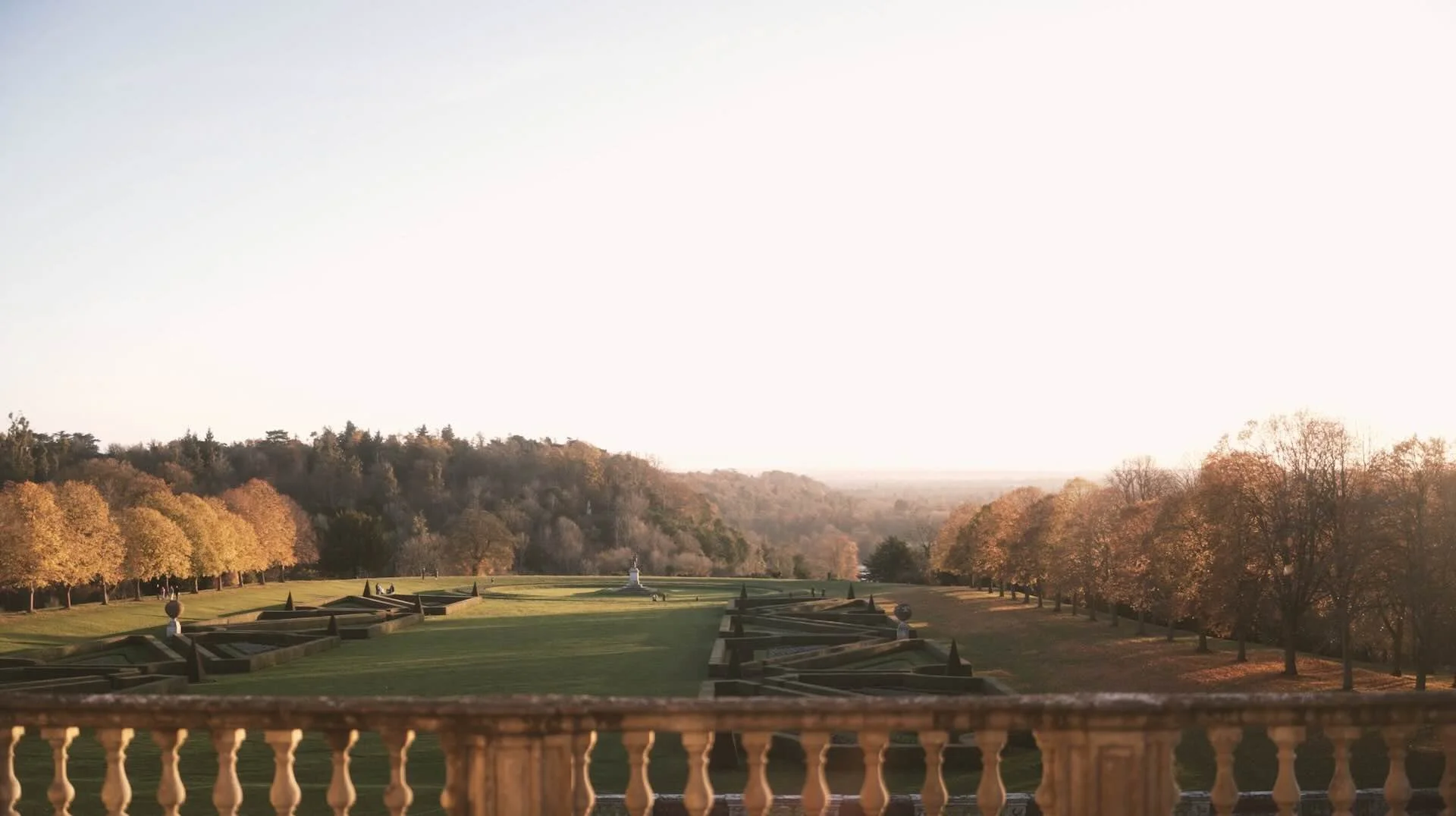 A landscaped garden with a row of trees and trimmed bushes, a fountain in the center, and a distant hilly landscape with trees during sunset.