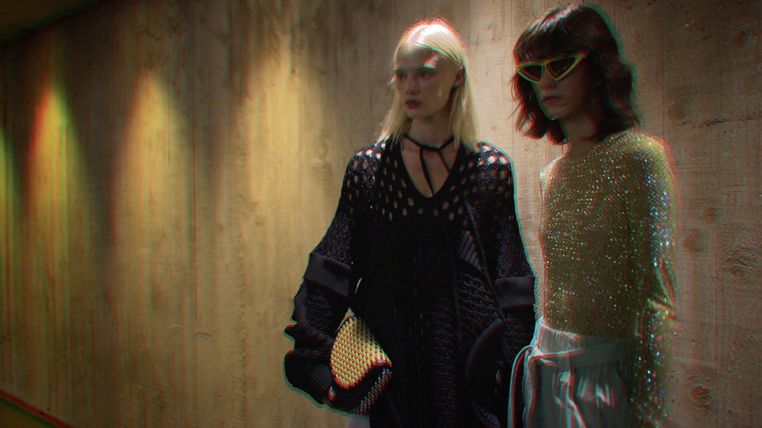 Two women with colorful hair and futuristic sunglasses standing against a wooden wall with kaleidoscopic lighting and effects.
