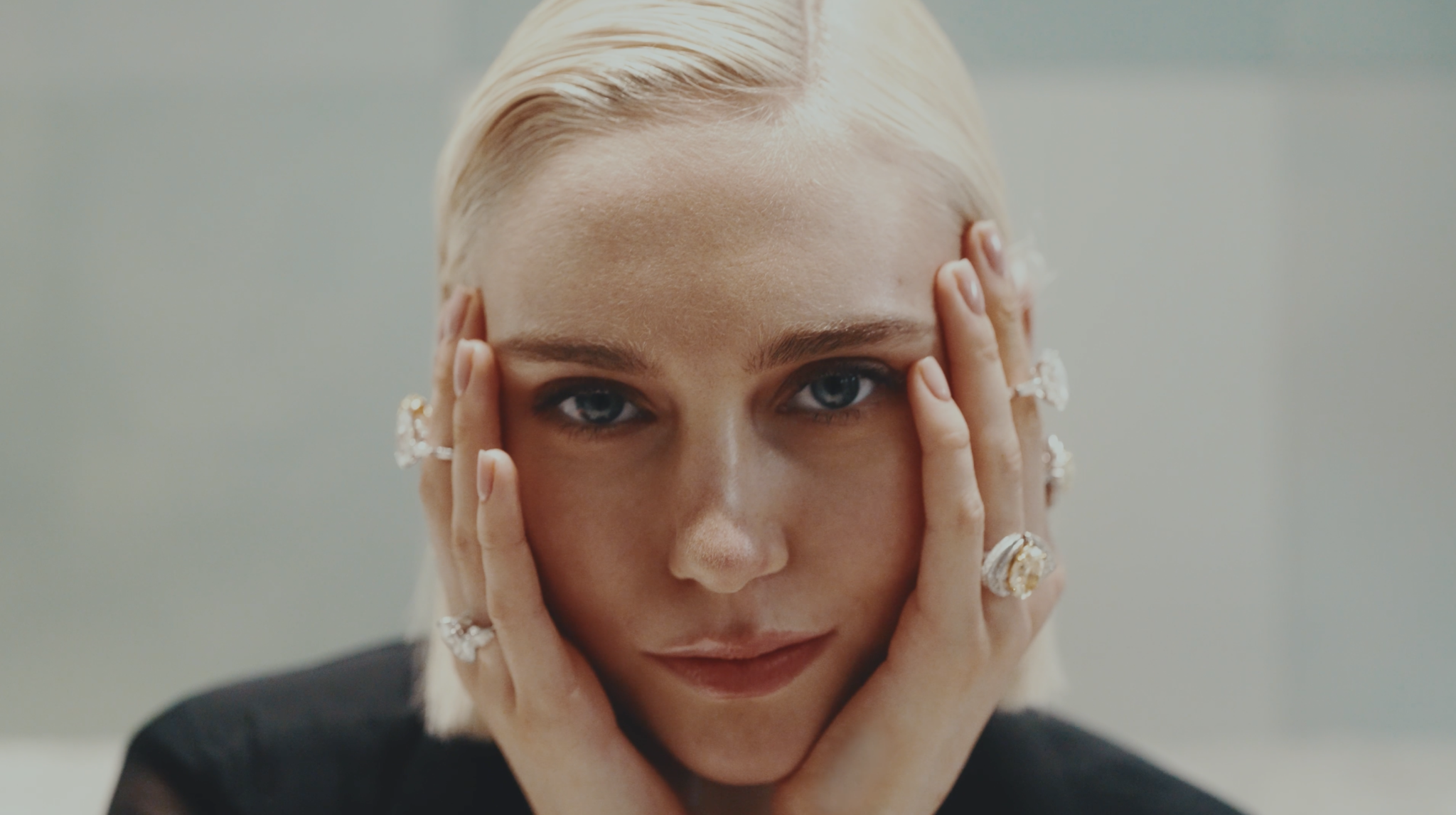 A close-up of a woman with platinum blonde hair, blue eyes, and multiple rings on her fingers, holding her face with both hands.