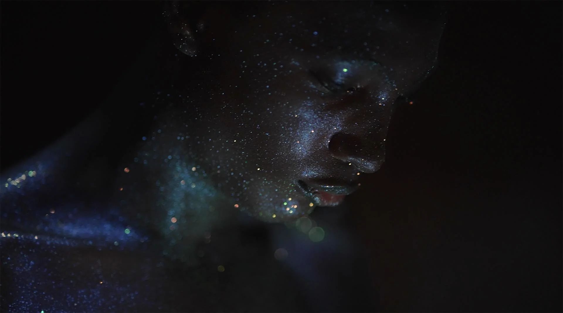 Close-up of a person's face covered in shimmering glitter, with their eyes closed, set against a dark background.