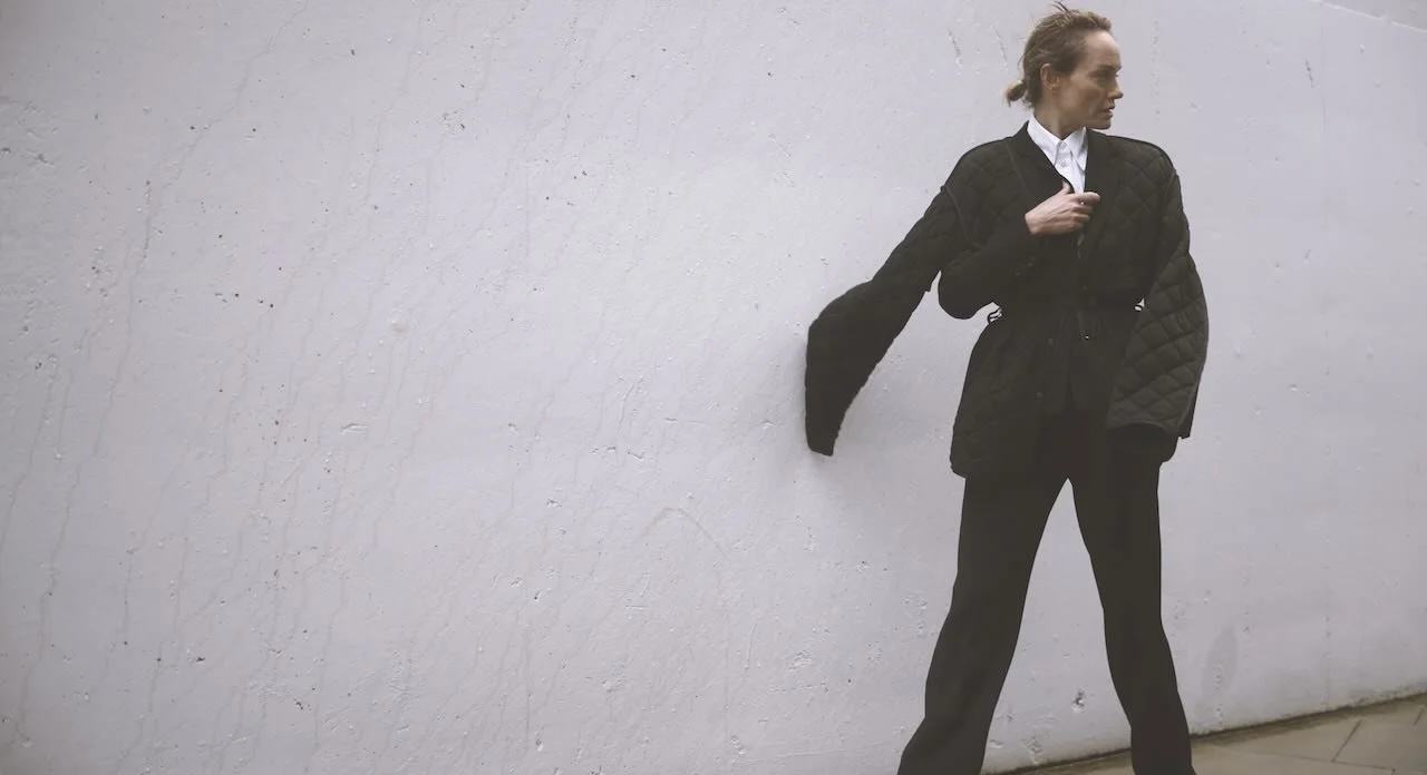 A woman standing against a plain white wall, wearing a black quilted jacket, white shirt, and black pants, looking to her right.