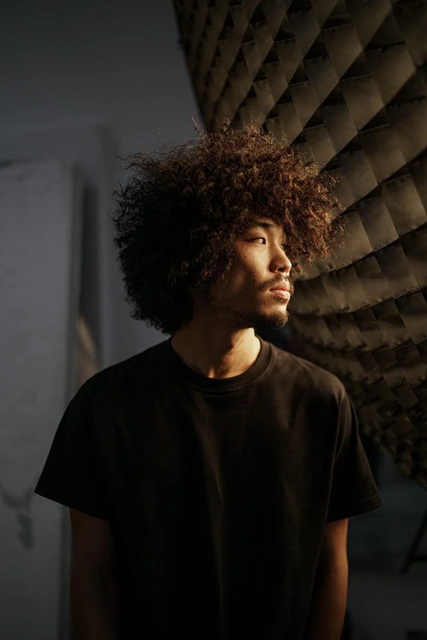 A young man with curly hair gazing thoughtfully to the right in a dark studio setting, with soundproofing foam on the wall beside him.