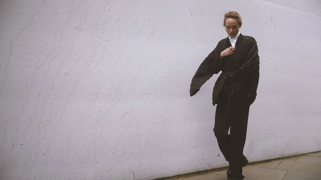 A woman stands against a plain, textured white wall, wearing a black quilted jacket over a white shirt, black pants, and black shoes.