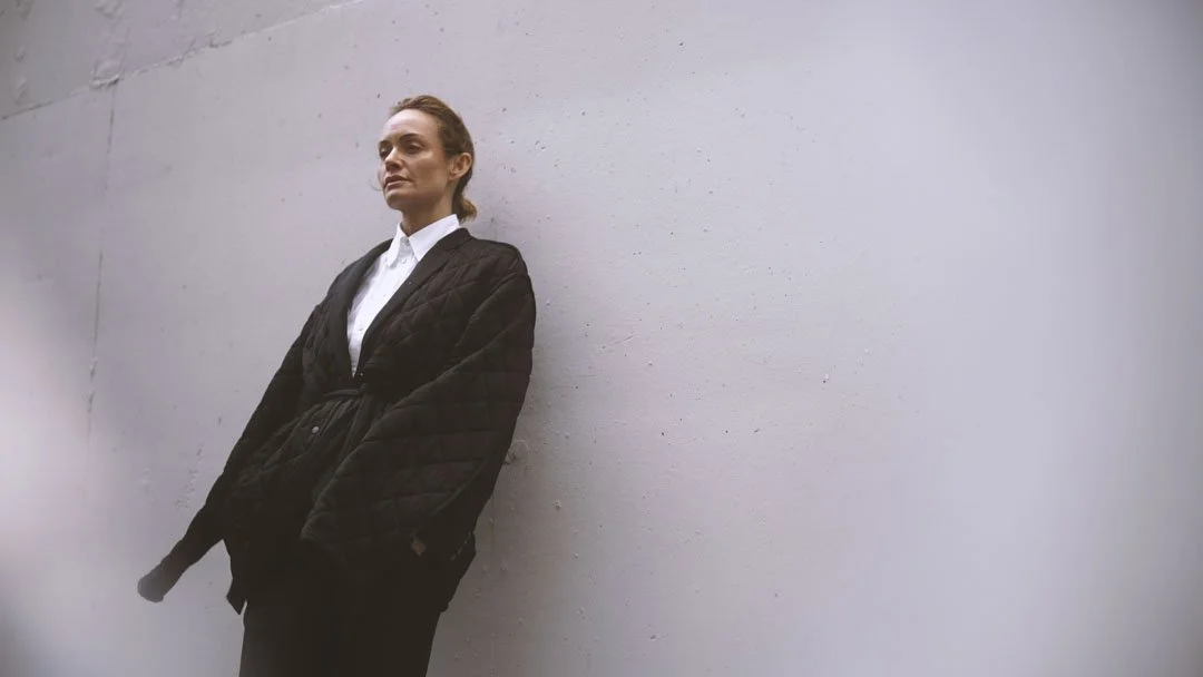 A woman leaning against a plain white wall, wearing a black quilted jacket and white shirt, with red hair pulled back.