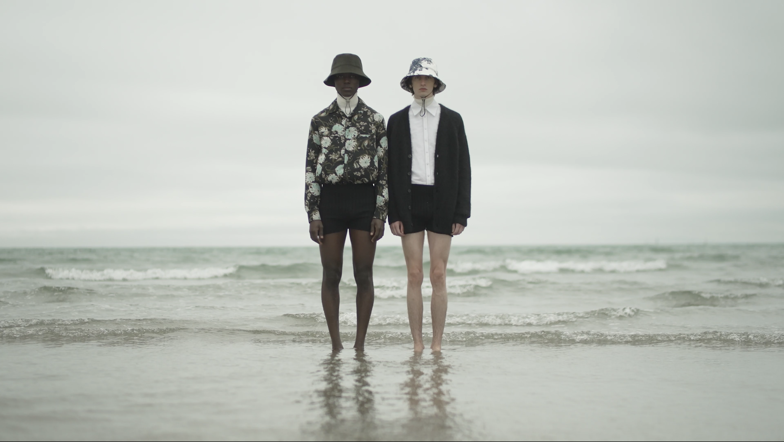 Two women standing in shallow ocean water, facing the camera, wearing hats and fashionable clothing, with overcast sky in the background.