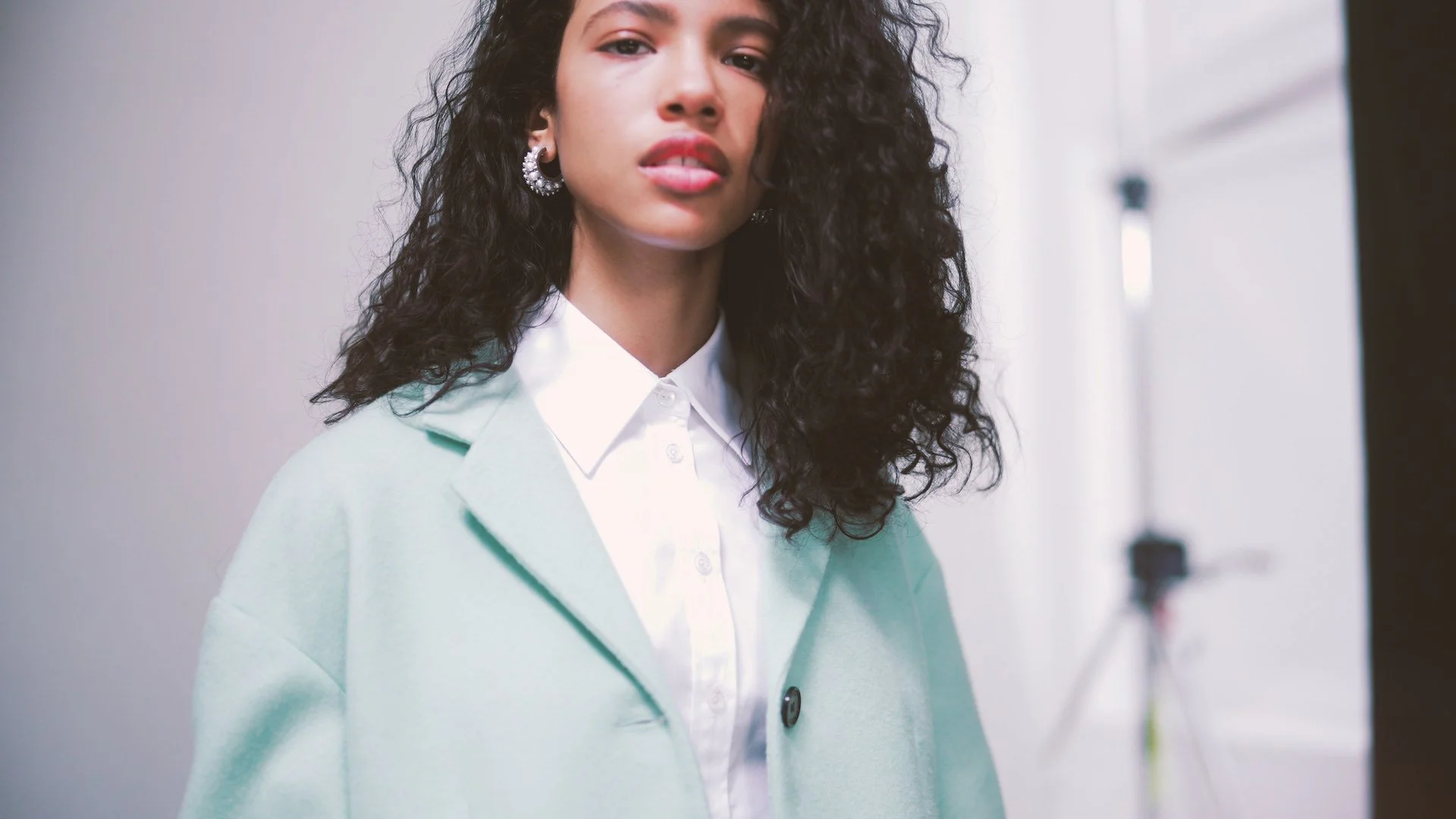 A young woman with curly black hair wearing a white collared shirt and a light green coat, standing indoors near a plain wall.