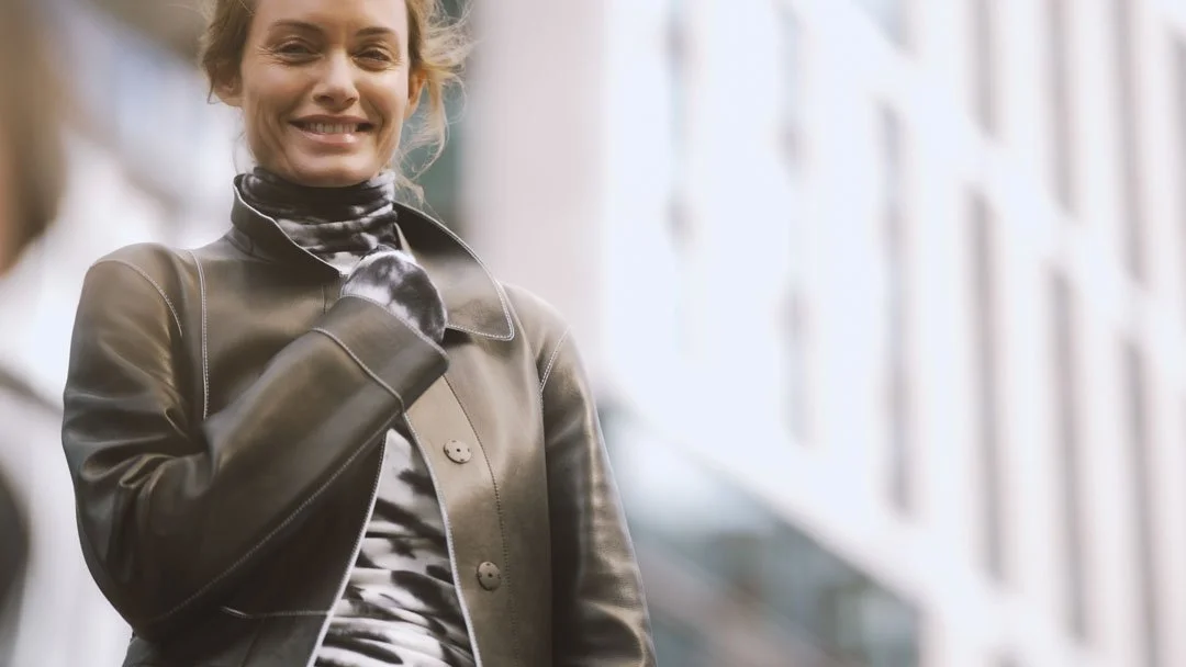 A smiling woman in a leather jacket and patterned scarf stands outdoors near a modern building with large windows.