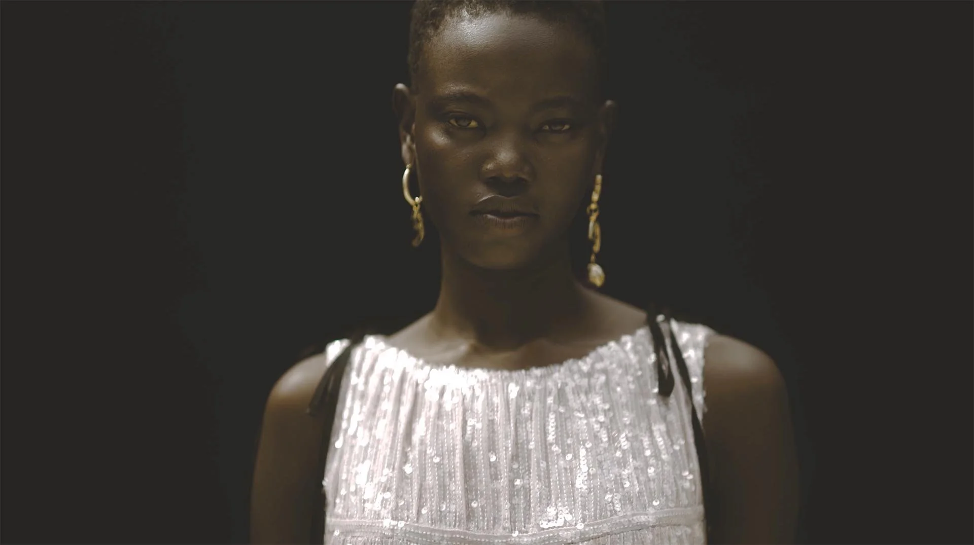A woman with dark skin and short hair wearing gold earrings and a shiny, silver dress with black straps, standing against a black background.