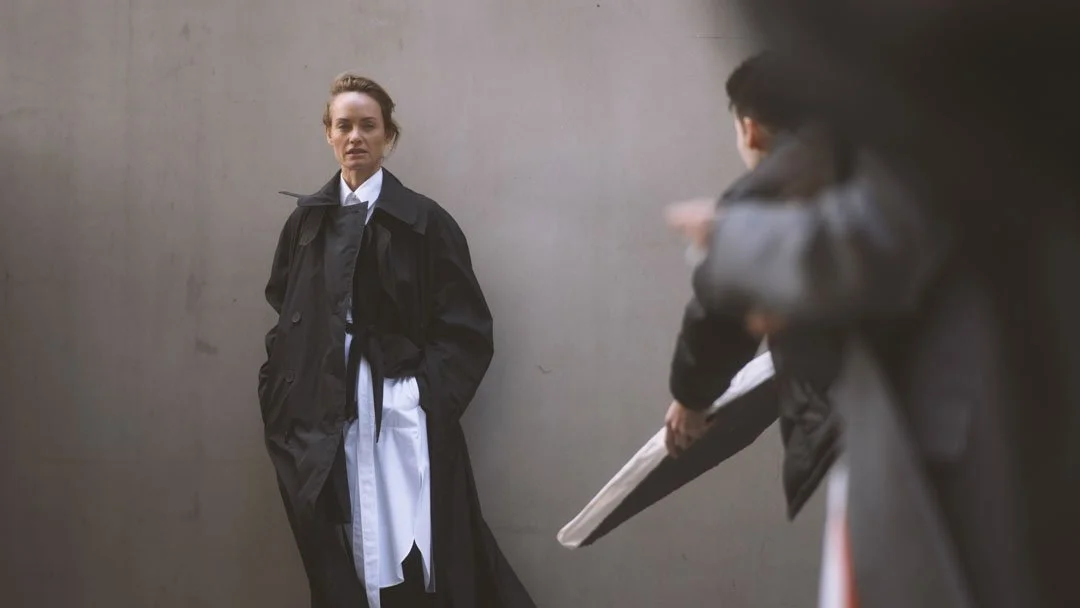 A woman standing against a plain wall while a photographer takes her picture.