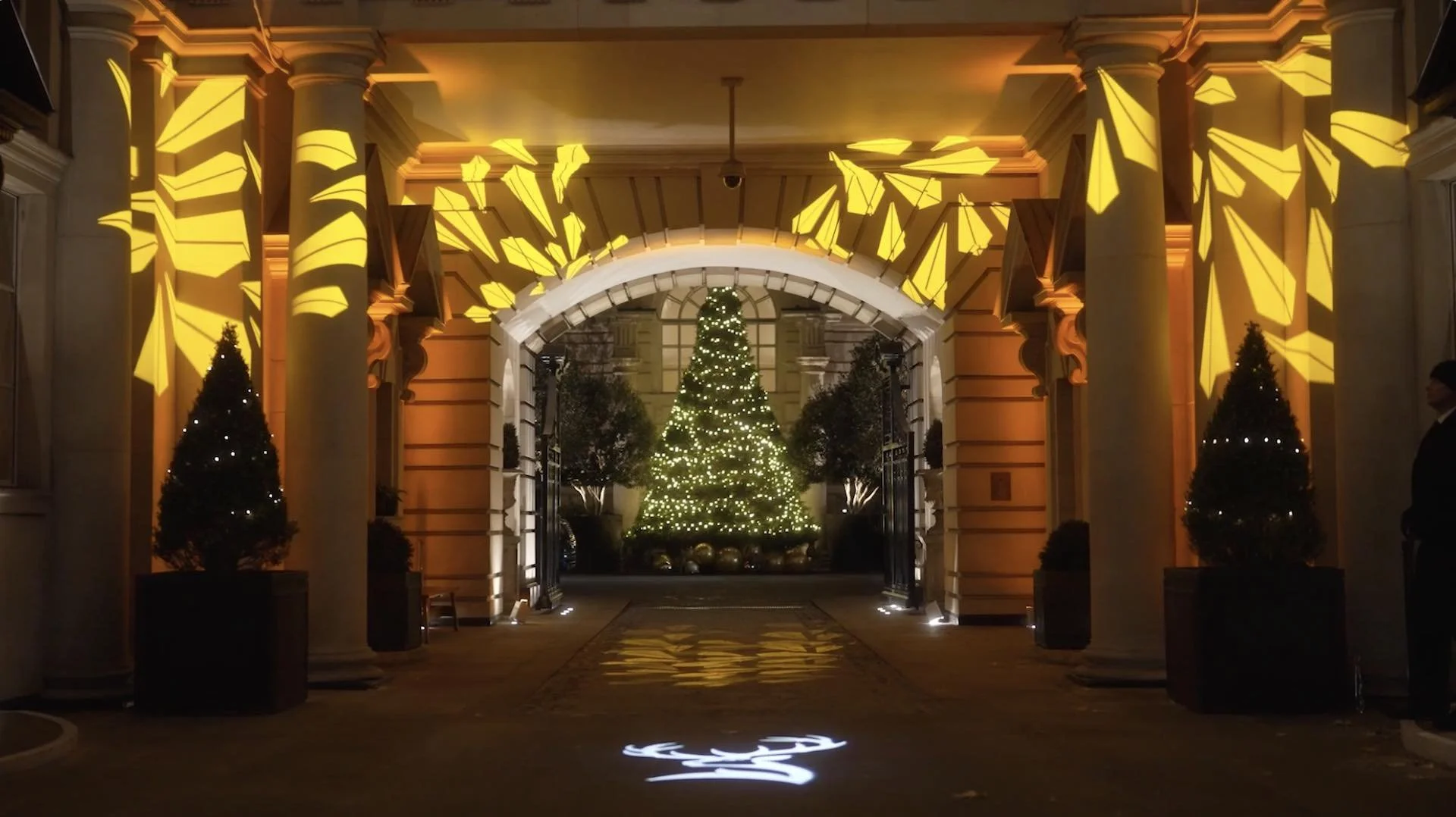 A decorated Christmas tree with white lights, centered in a grand entryway with tall white columns and warm yellow lighting. Smaller trees with lights flank the entrance, and yellow geometric light projections are on the ceiling and columns.