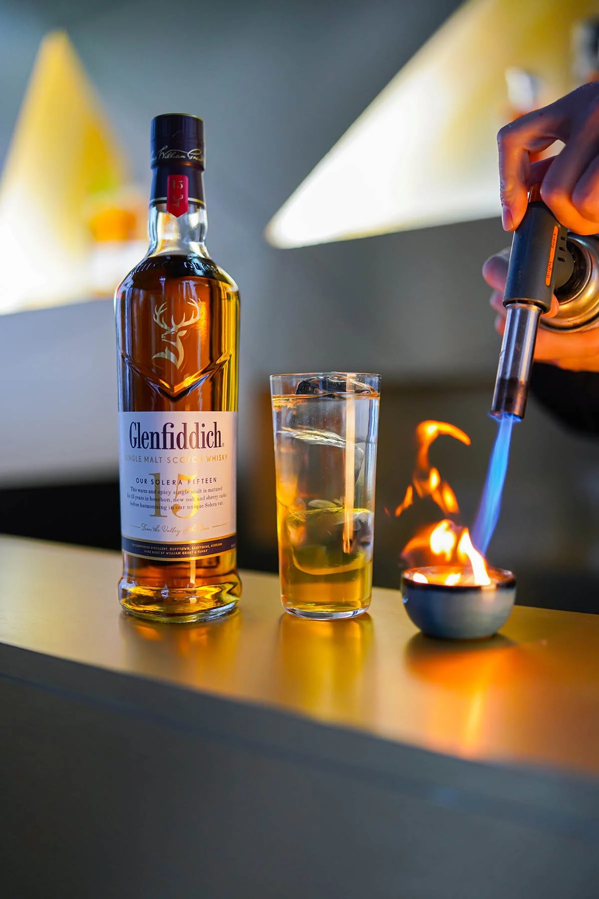 A bottle of Glenfiddich single malt Scotch whisky, a glass filled with whisky and ice, and a person using a flamethrower on a small dish, all on a yellow surface with a blurred background.