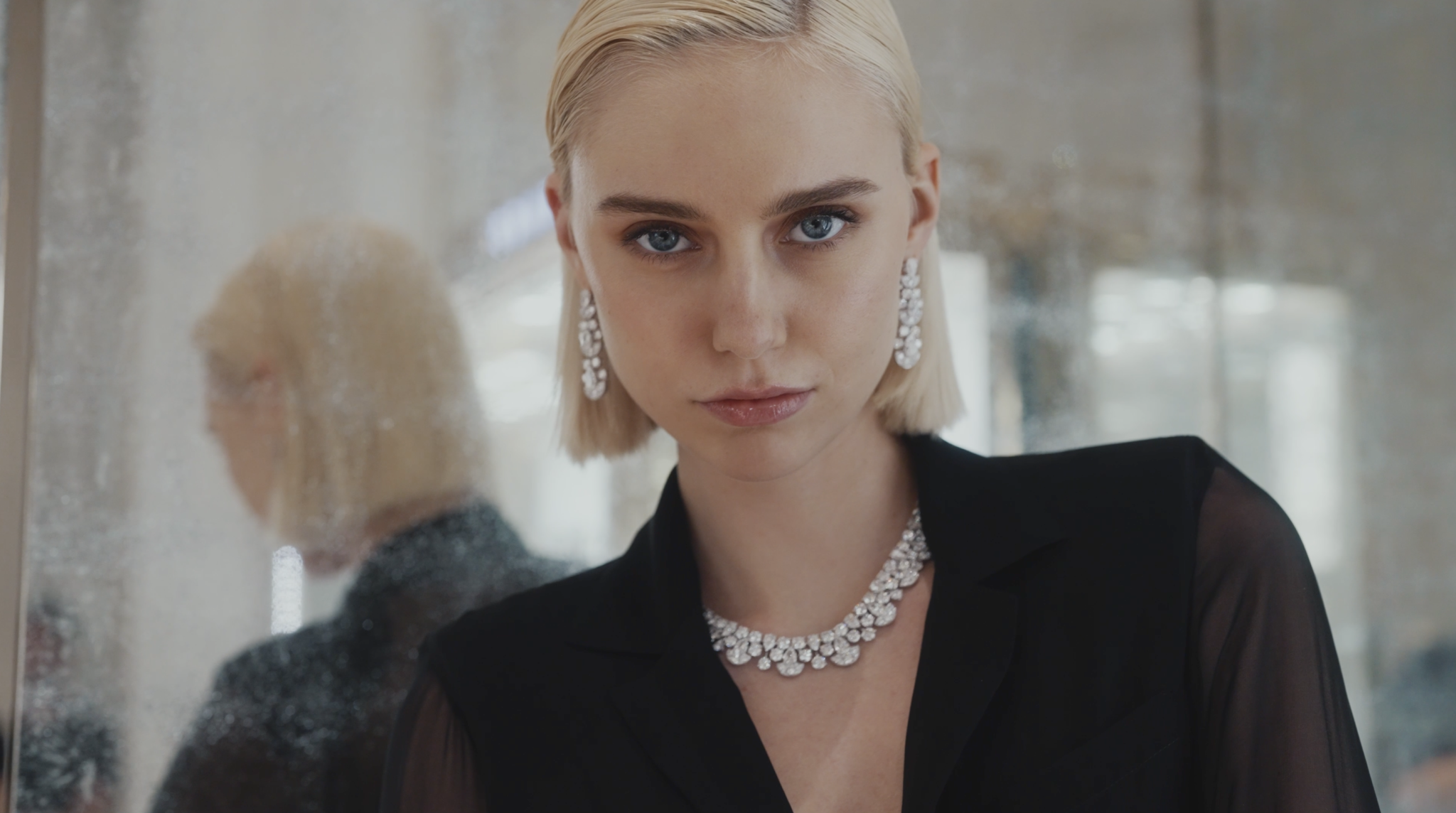 Close-up of a young woman with short platinum blonde hair and blue eyes, wearing diamond jewelry including earrings and a necklace, dressed in a black top, looking directly at the camera with a neutral expression.