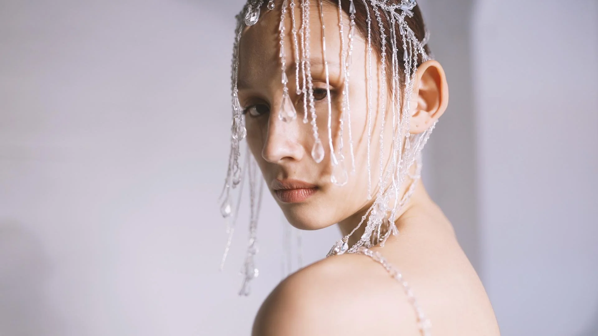 A woman with wet hair and bare shoulders, wearing a decorative bead and crystal headpiece with matching jewelry, looking over her shoulder against a plain background.