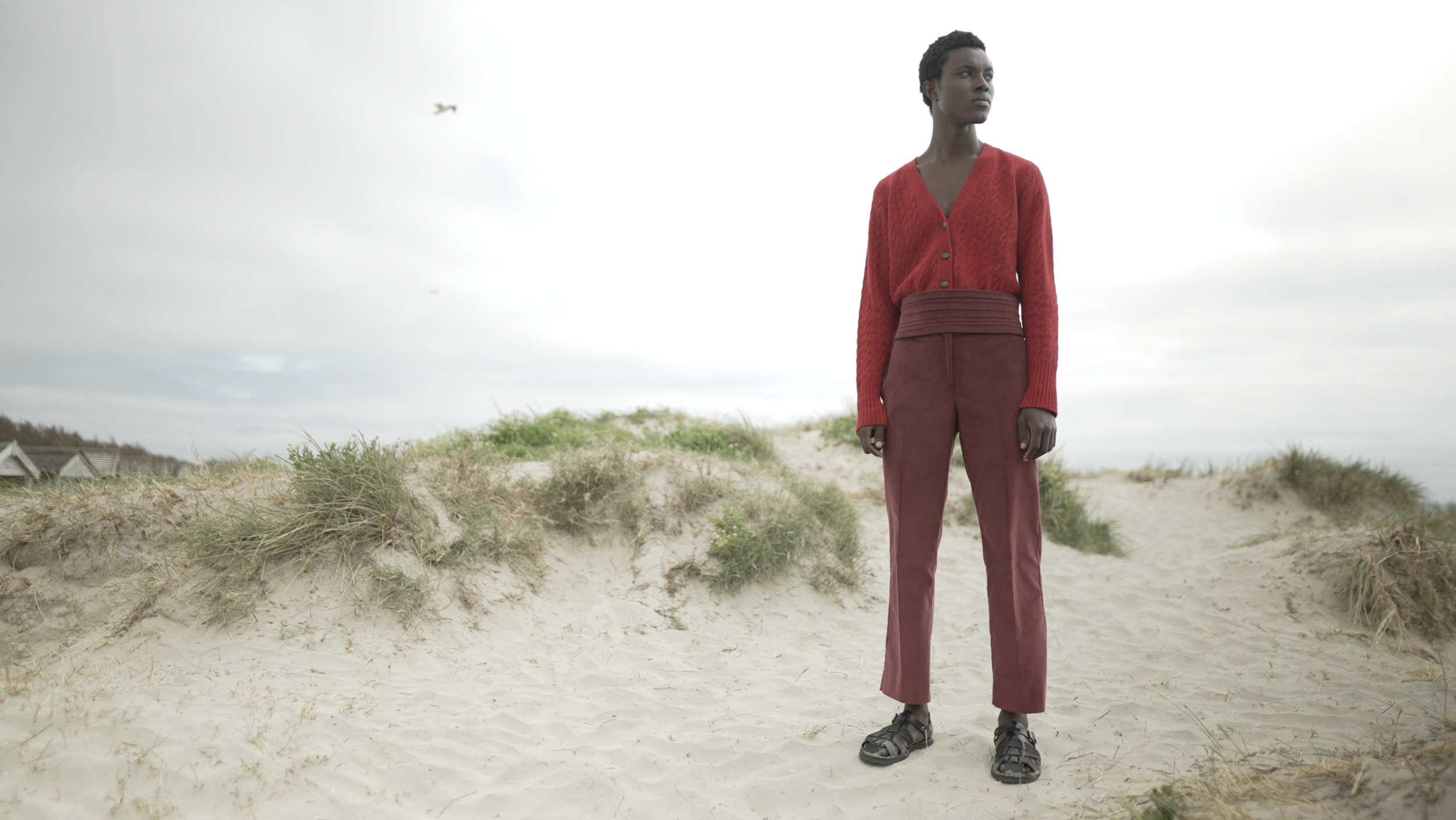 A person in a red cardigan and red pants standing on sandy dunes with grasses and a cloudy sky in the background.