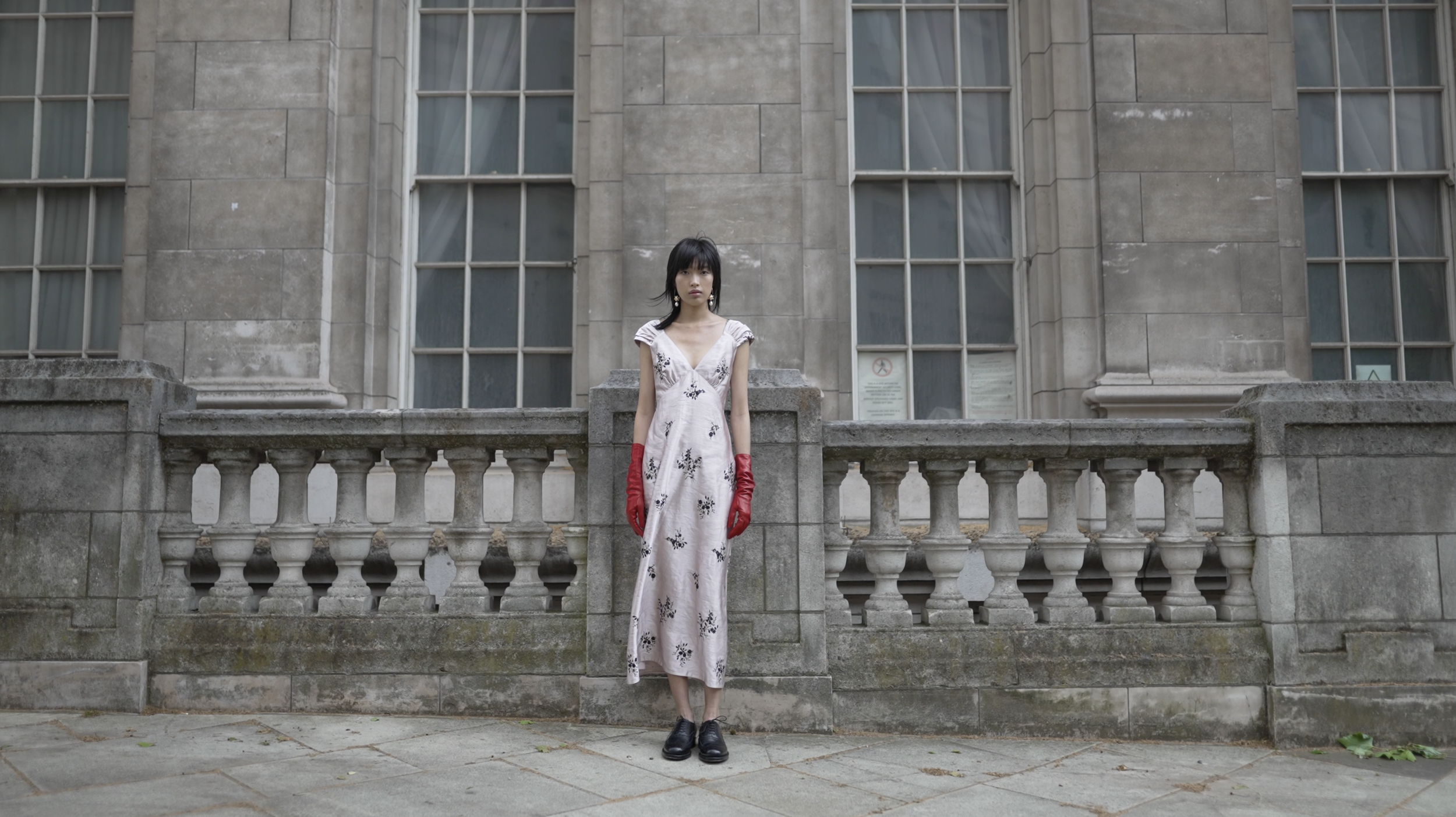 A woman wearing a white floral dress, black shoes, and red gloves standing on a stone terrace in front of a historic building with large windows and stone balustrades.