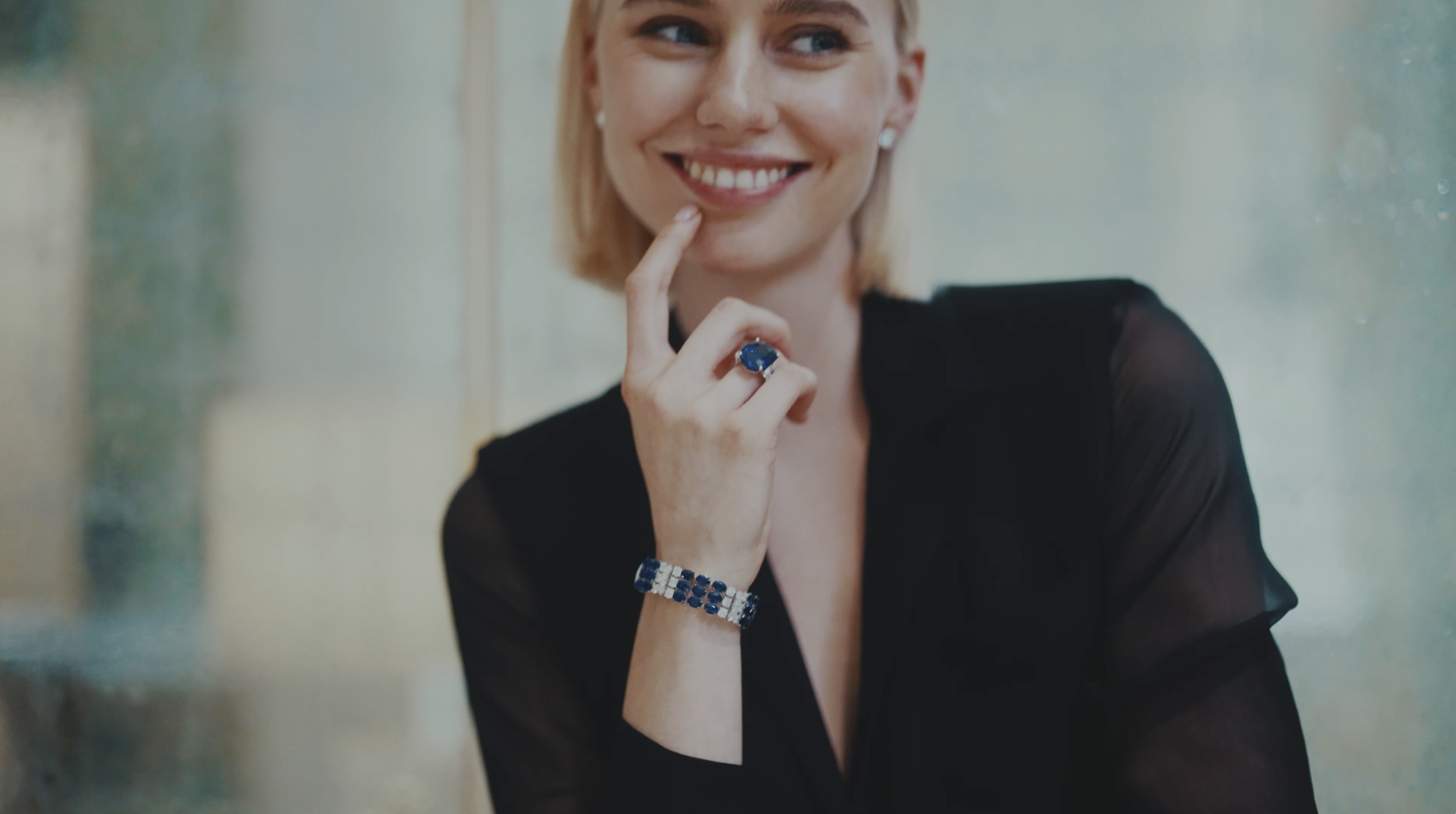 Smiling woman with short blonde hair wearing a black sheer top, a large blue ring, and matching blue jewelry, posing indoors near a reflective surface.