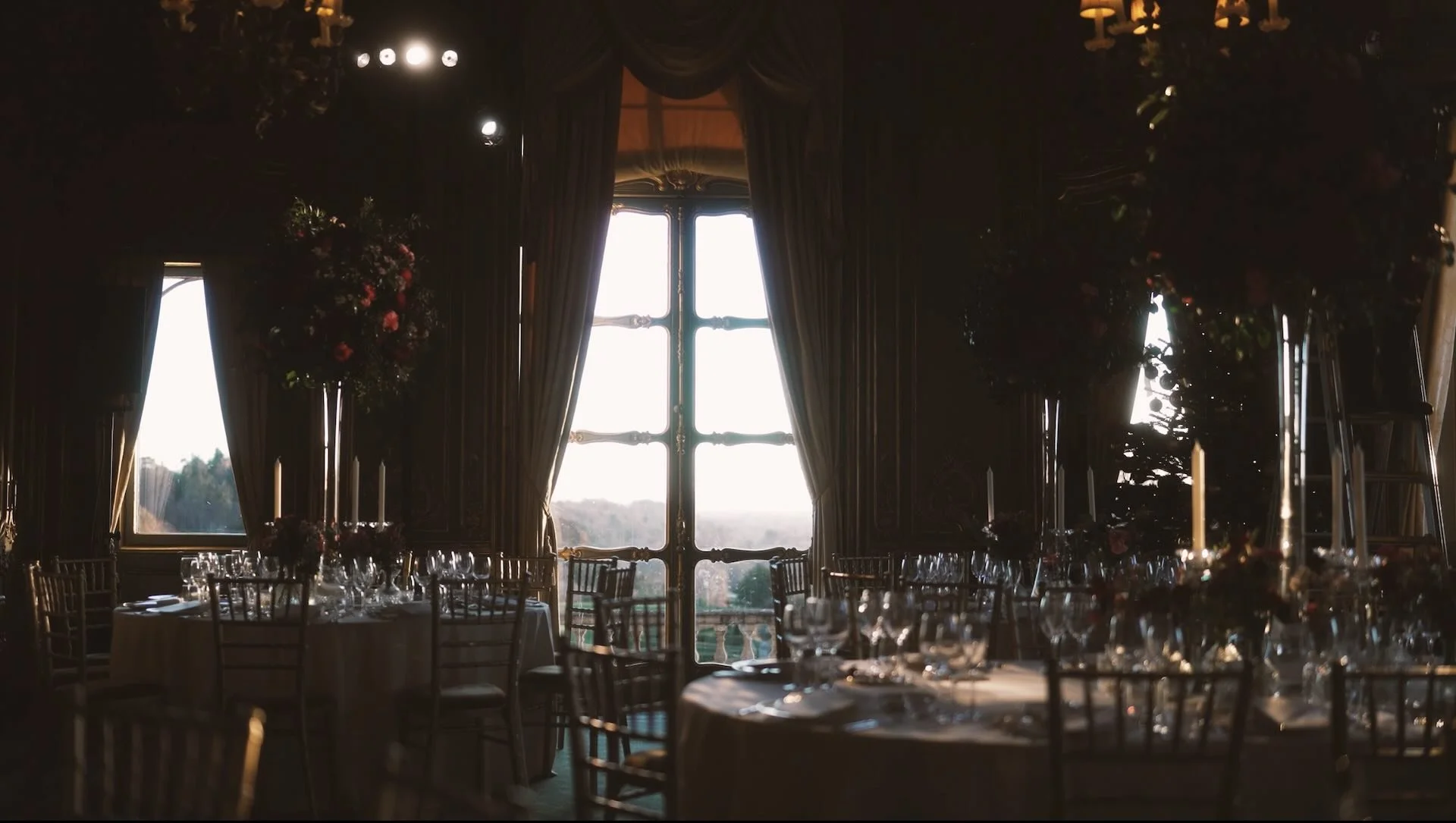 A dimly lit elegant banquet hall with large windows draped in dark curtains, decorated with floral arrangements and set tables with wine glasses, candles, and chairs, likely for a formal event or wedding reception.