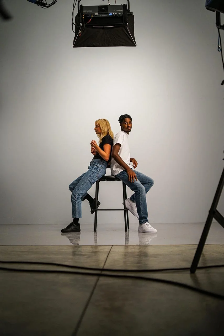 Two people sitting back-to-back on a stool in a photography studio with a plain white background and studio equipment overhead.