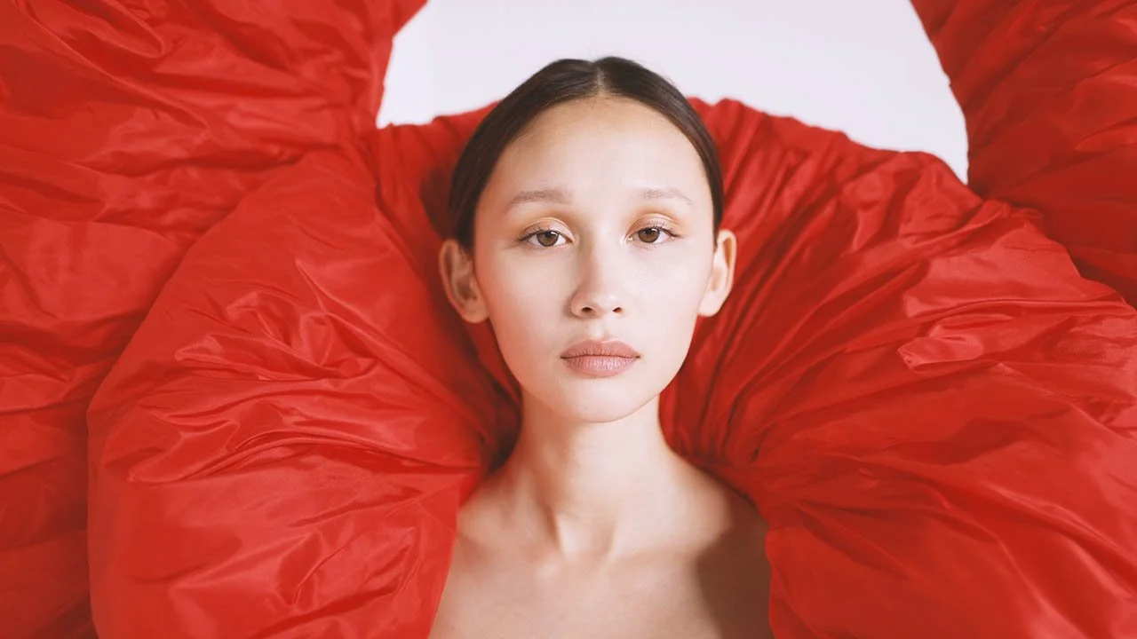 Close-up of a woman with fair skin and dark hair, surrounded by large red fabric leaves or petals, creating an artistic and elegant portrait.