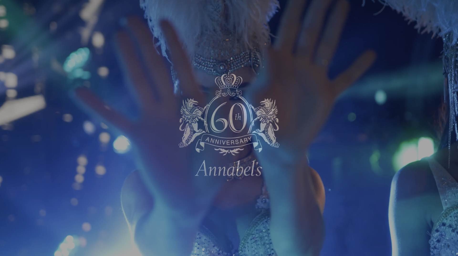 Celebrating Annabel's 60th anniversary with a person standing behind a glass window that has the anniversary logo, wearing ornate jewelry and a fancy outfit, with colorful lights in the background.