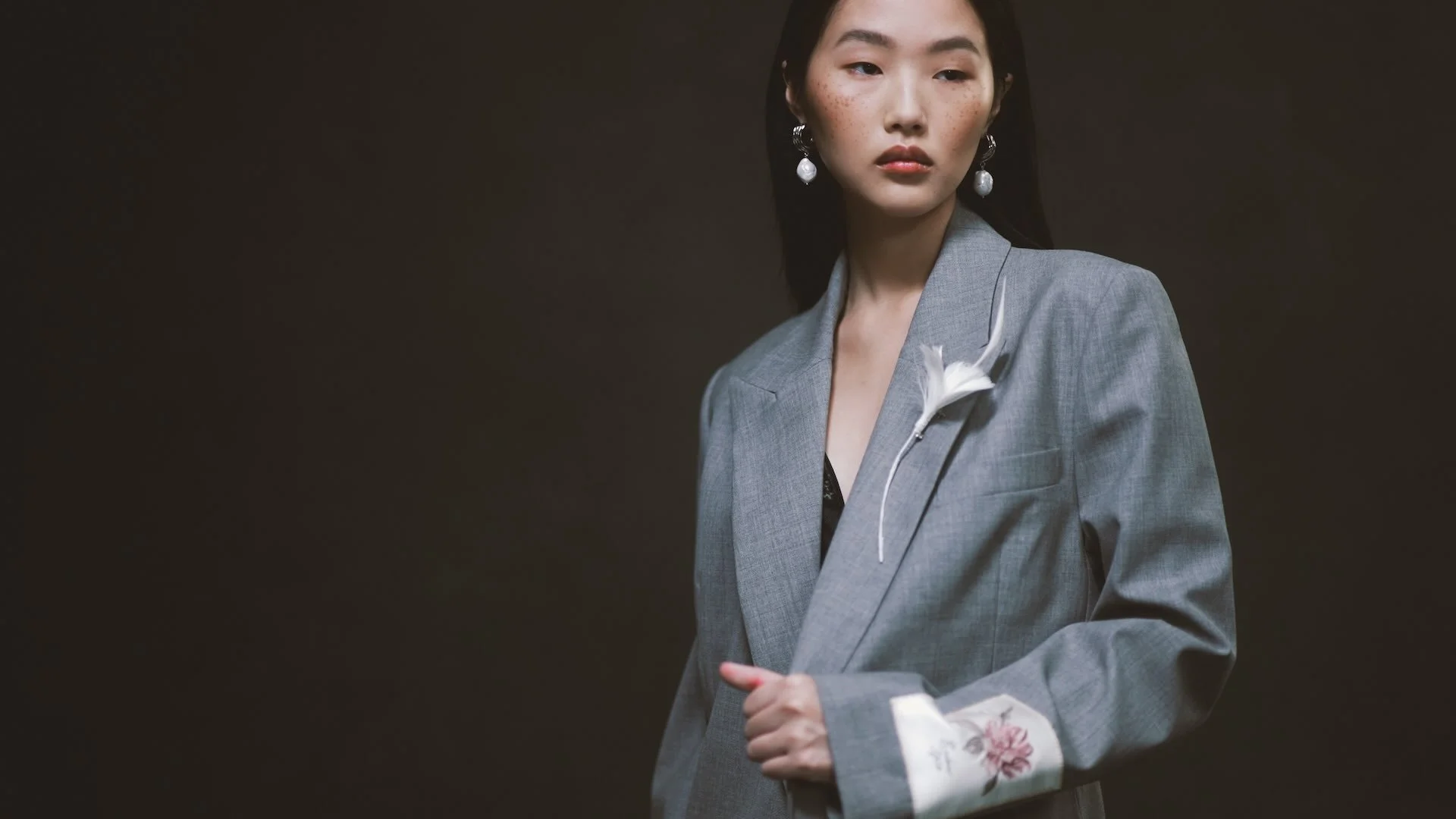 A woman with black hair, wearing a gray blazer with floral embroidered cuffs and large pearl earrings, holding a white flower with gingerly clasped hands, against a dark background.