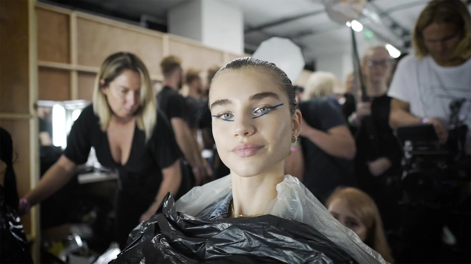 A young woman with elaborate makeup and styled hair sitting in a busy backstage area preparing for a photoshoot or fashion show.