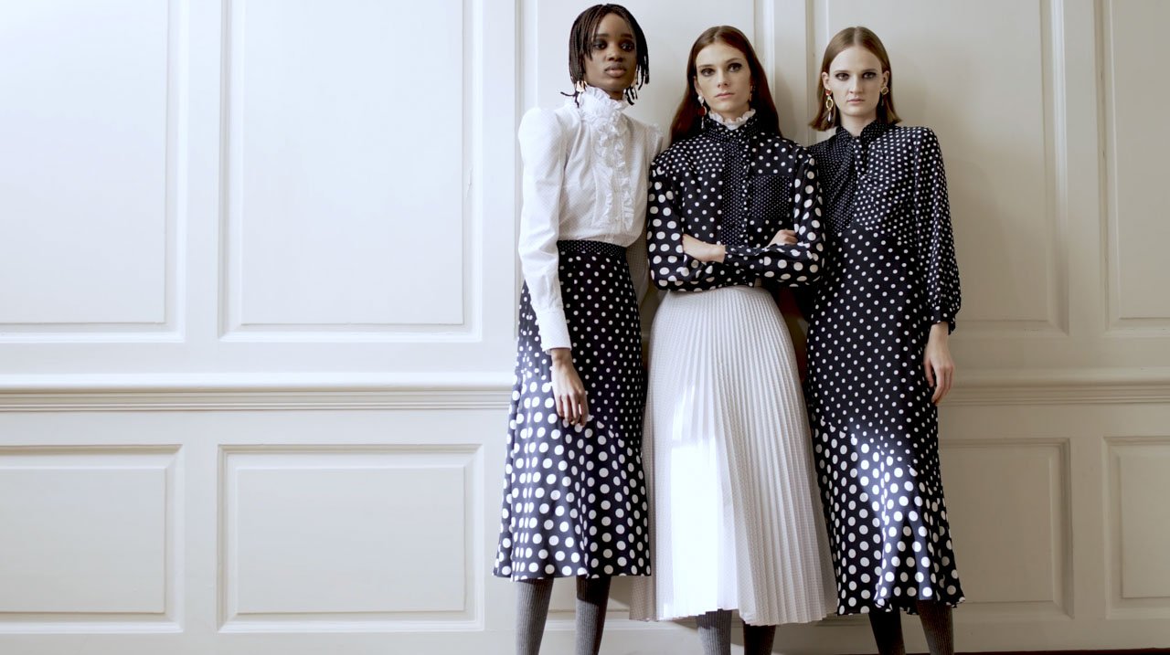 Three women standing in front of a white paneled wall, wearing black and white polka dot and white clothing, with serious expressions.
