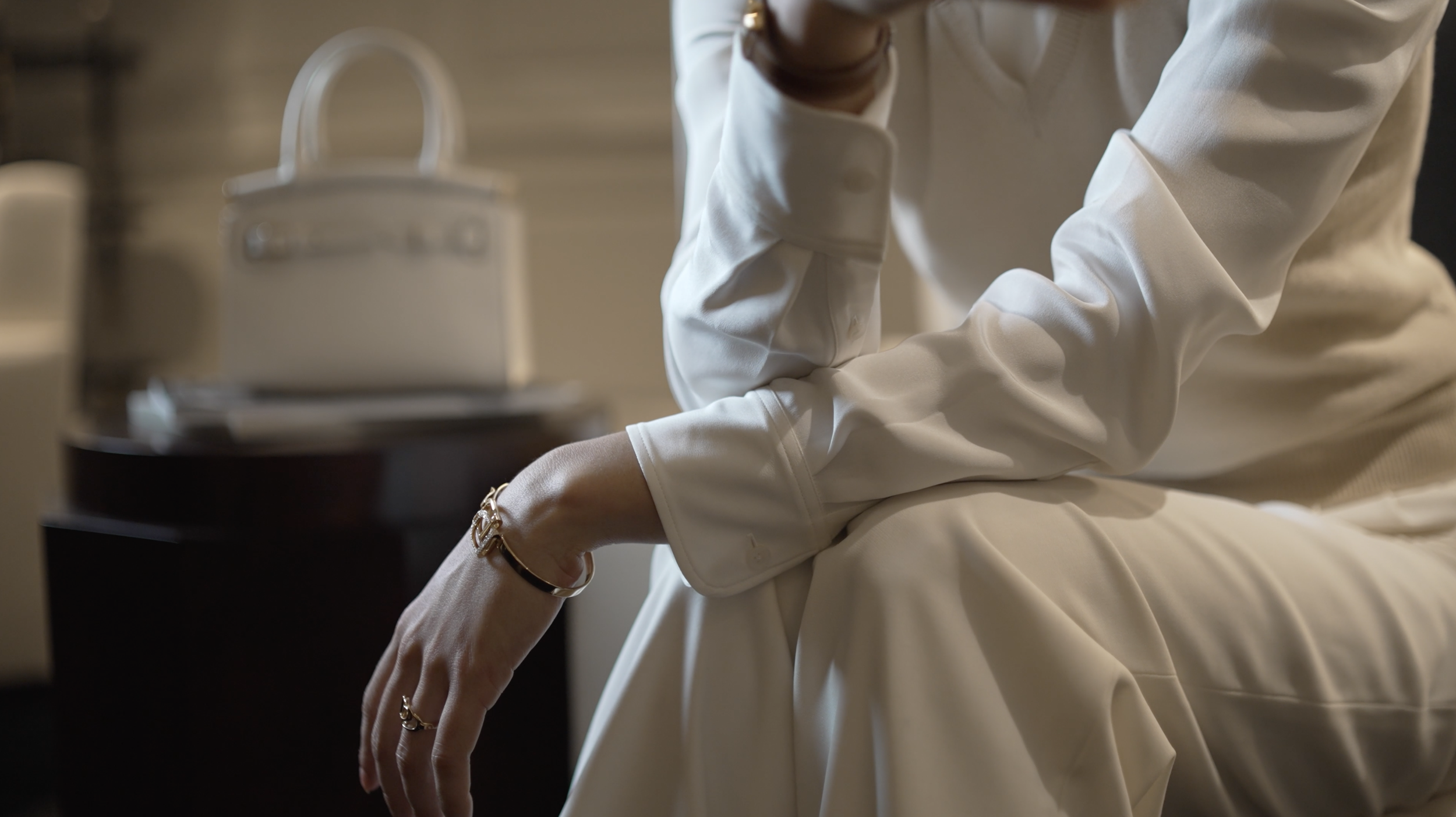 A person wearing a white blouse and beige pants, sitting with one hand resting on their knee and their other arm crossed, with a handbag and jewelry visible.