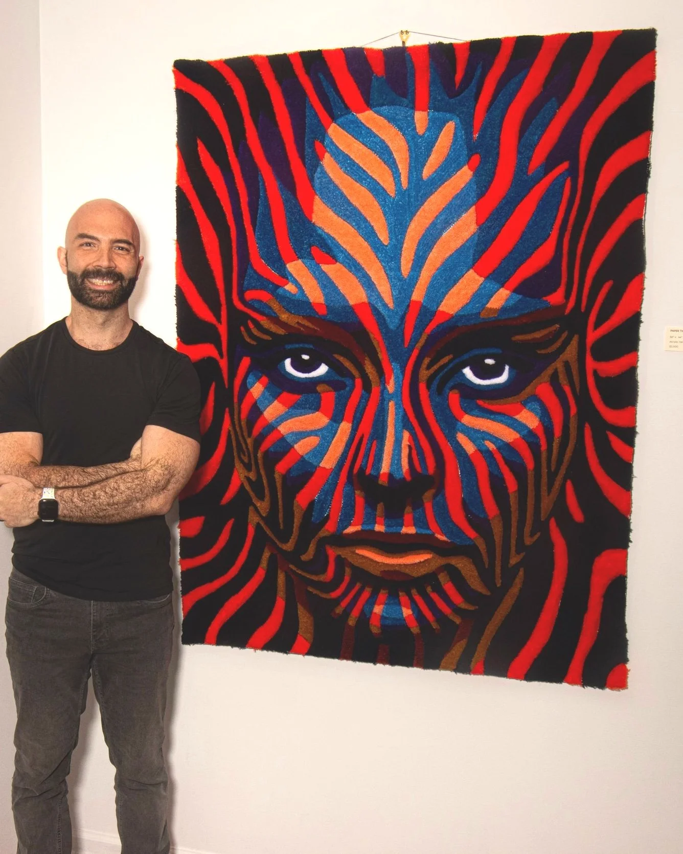 A man with a beard, wearing a black t-shirt and gray jeans, standing with his arms crossed in front of a colorful abstract art piece depicting a person's face with intense eyes, using bold lines and vibrant colors like red, blue, and brown.