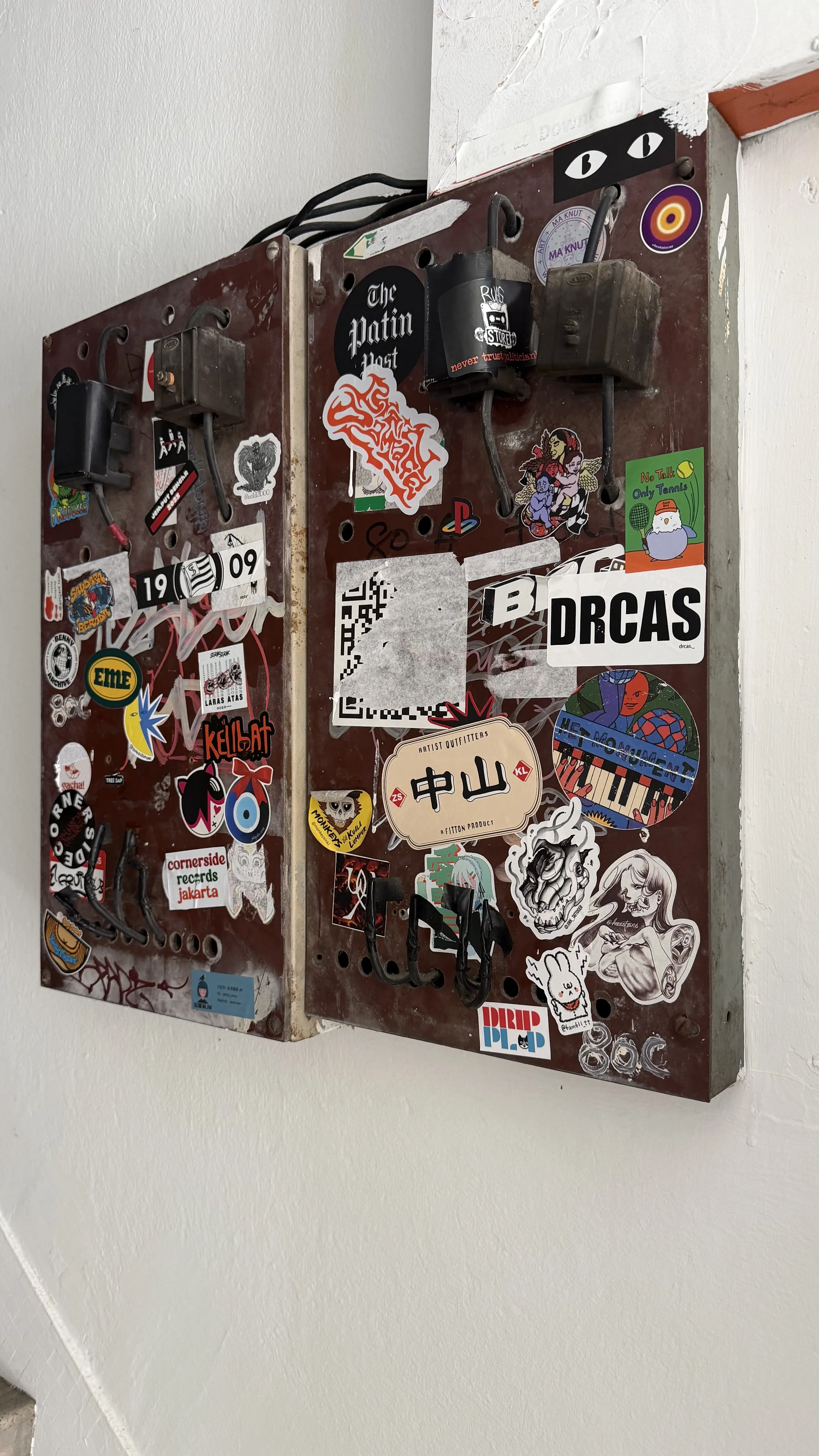 A mixed collection of various colorful stickers on a metal electrical box mounted on a white wall.