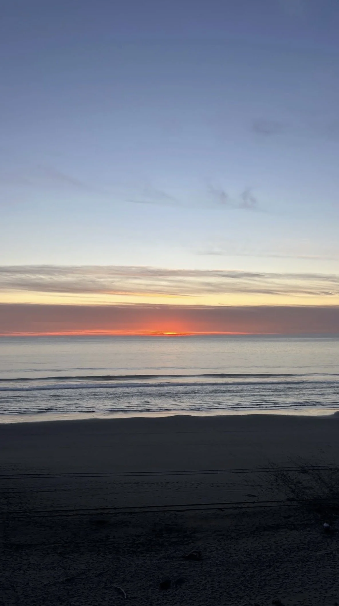 Beach with calm ocean waves at sunset, with a partly cloudy sky and dark sand in the foreground.