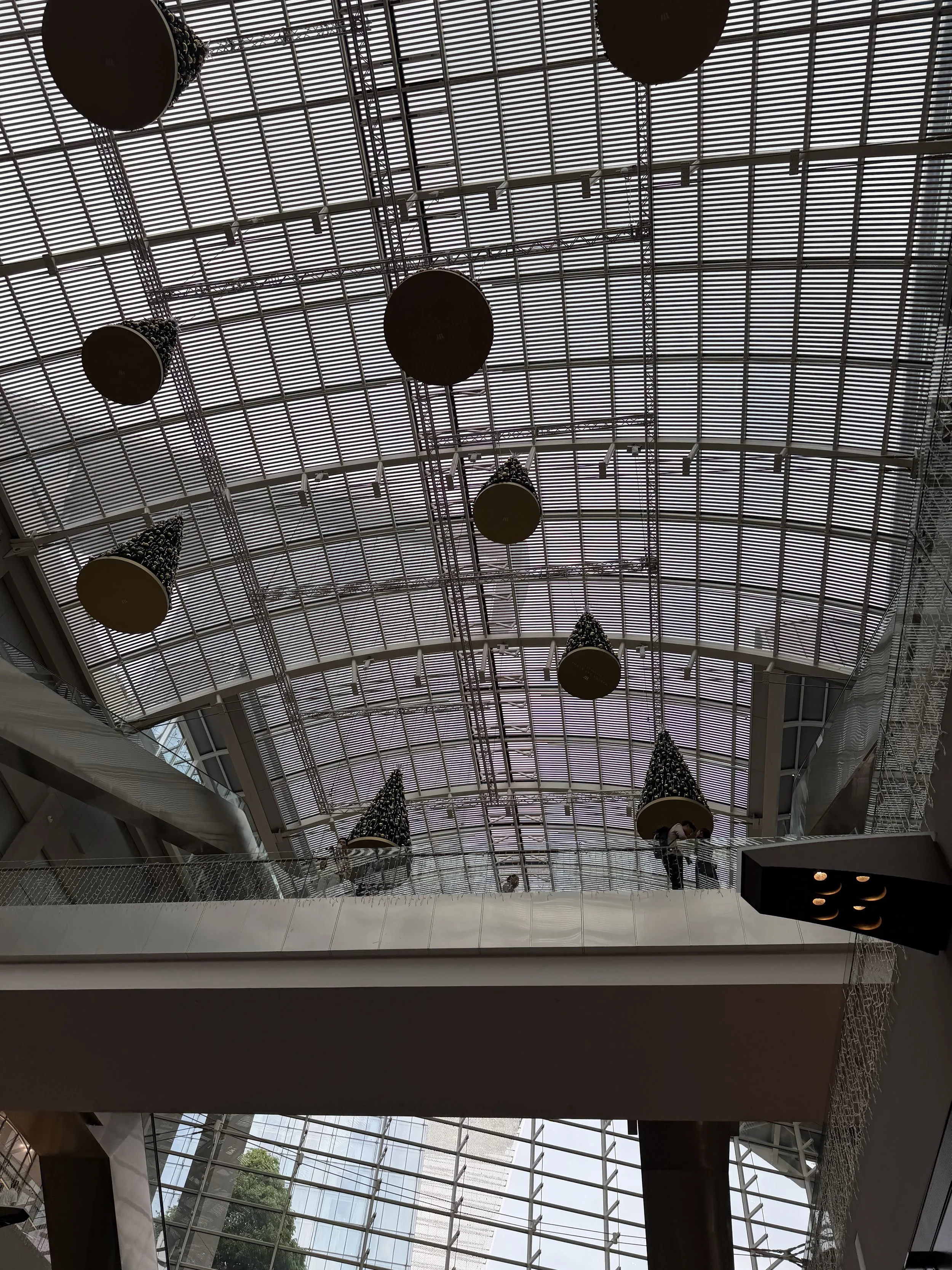 Inside of a modern shopping mall or airport with a high glass ceiling, pendant lights, and decorative conical Christmas trees.