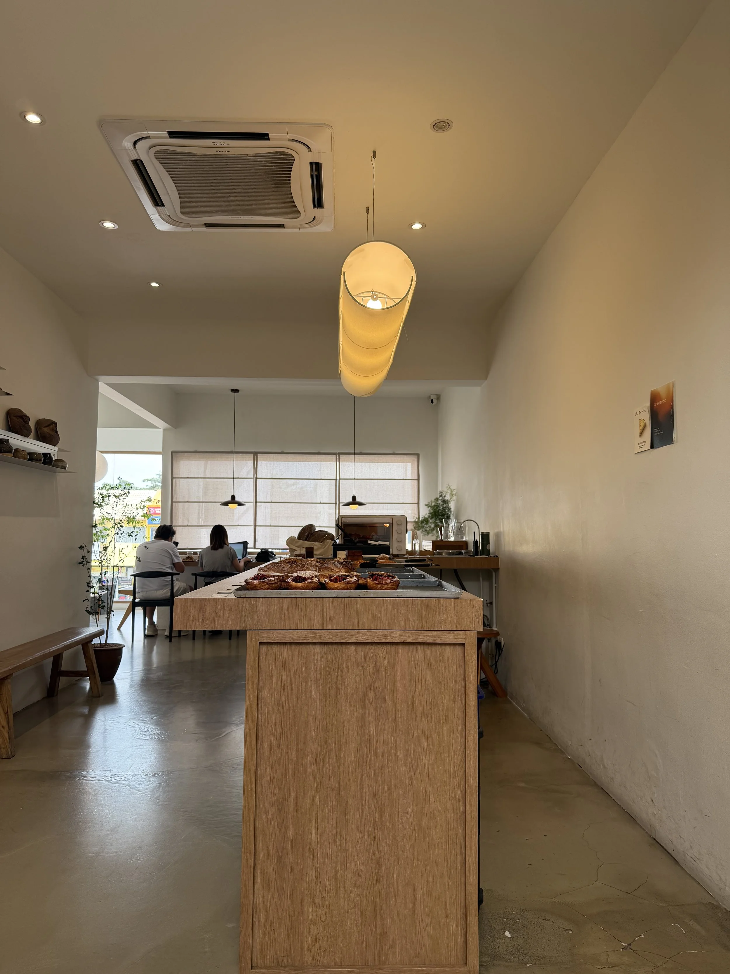 Interior of a cafe with a wooden table in the foreground, pastries on display, and customers seated at tables in the background near a window.