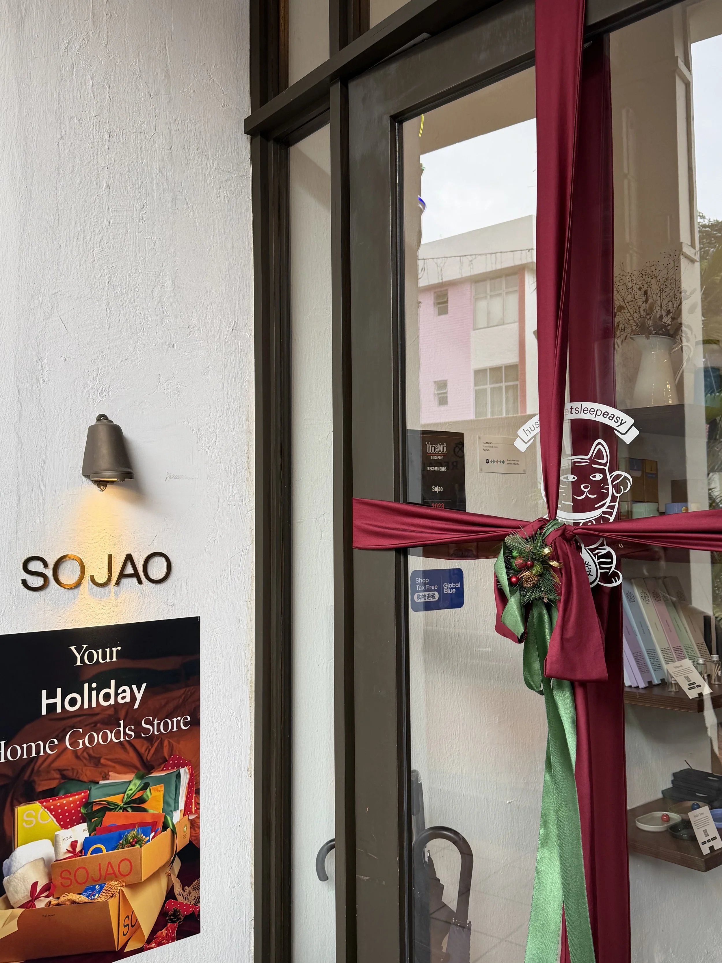 Decorated store entrance with red and green ribbon, holiday sign, and store sign 'SO JAO' on white wall.