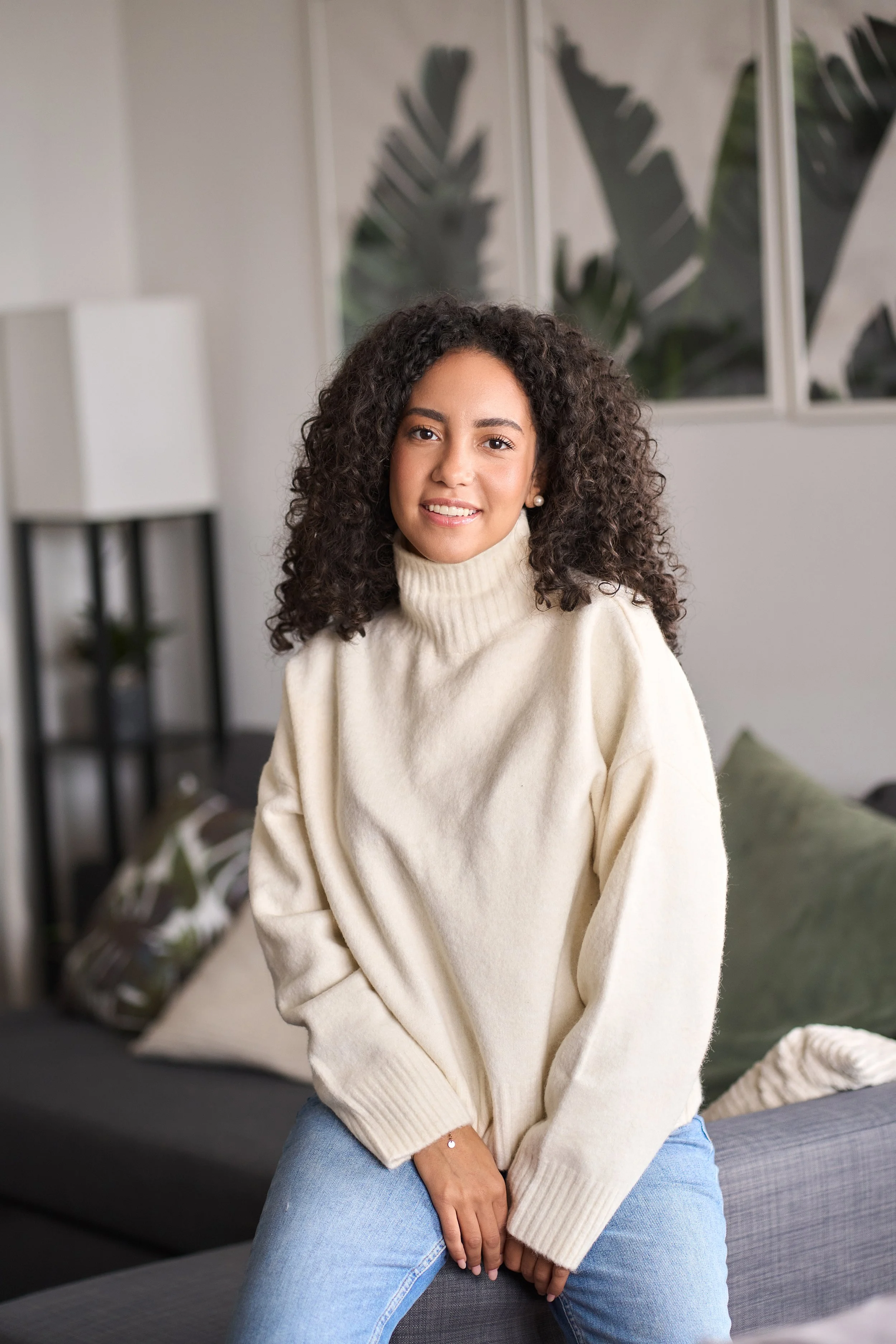 A woman with curly hair sitting on a gray couch in a living room with plants and windows in the background.