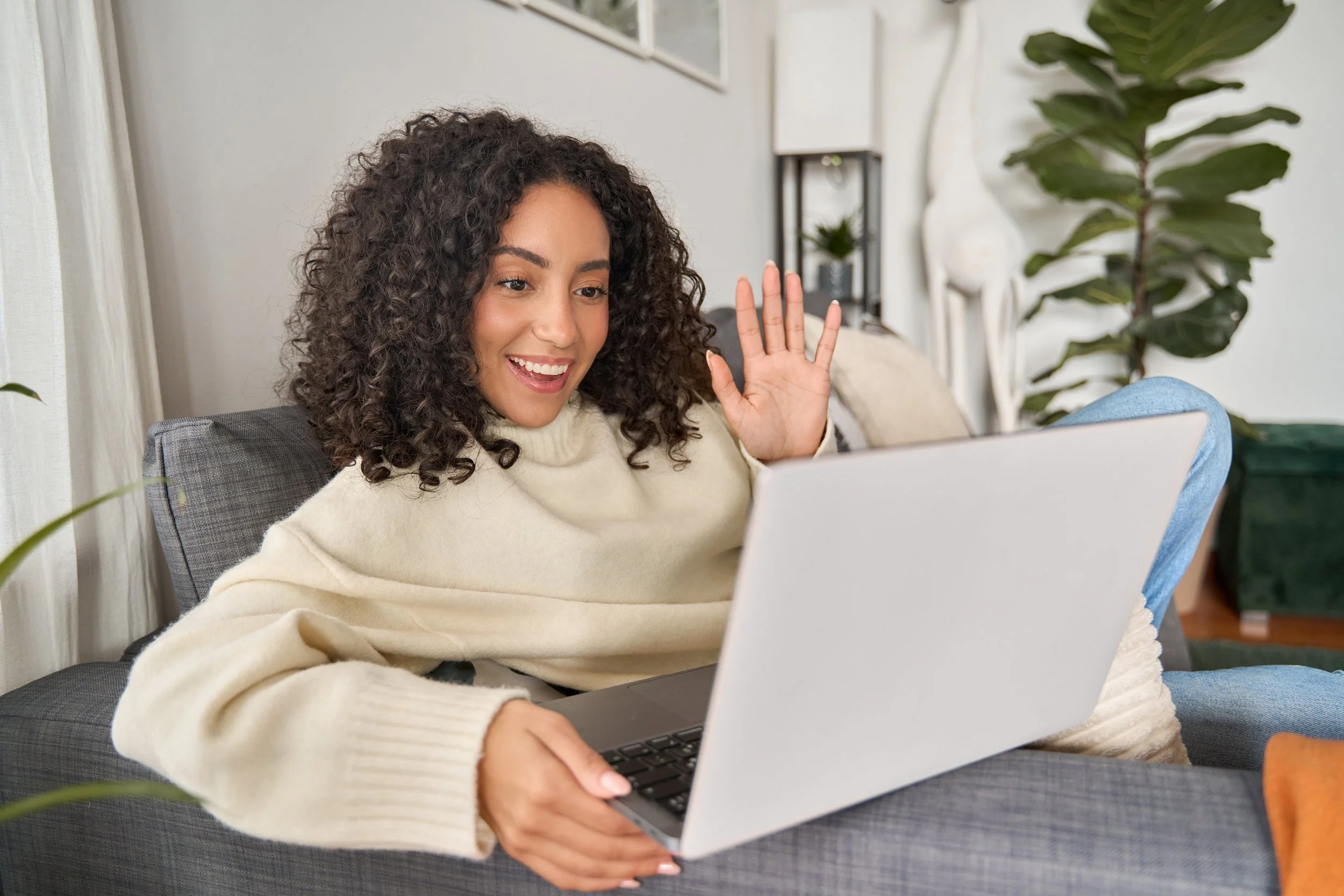 Young woman with curly hair sitting on a gray sofa, smiling and waving while using a silver laptop in a well-lit living room with large green plant and art on the wall.