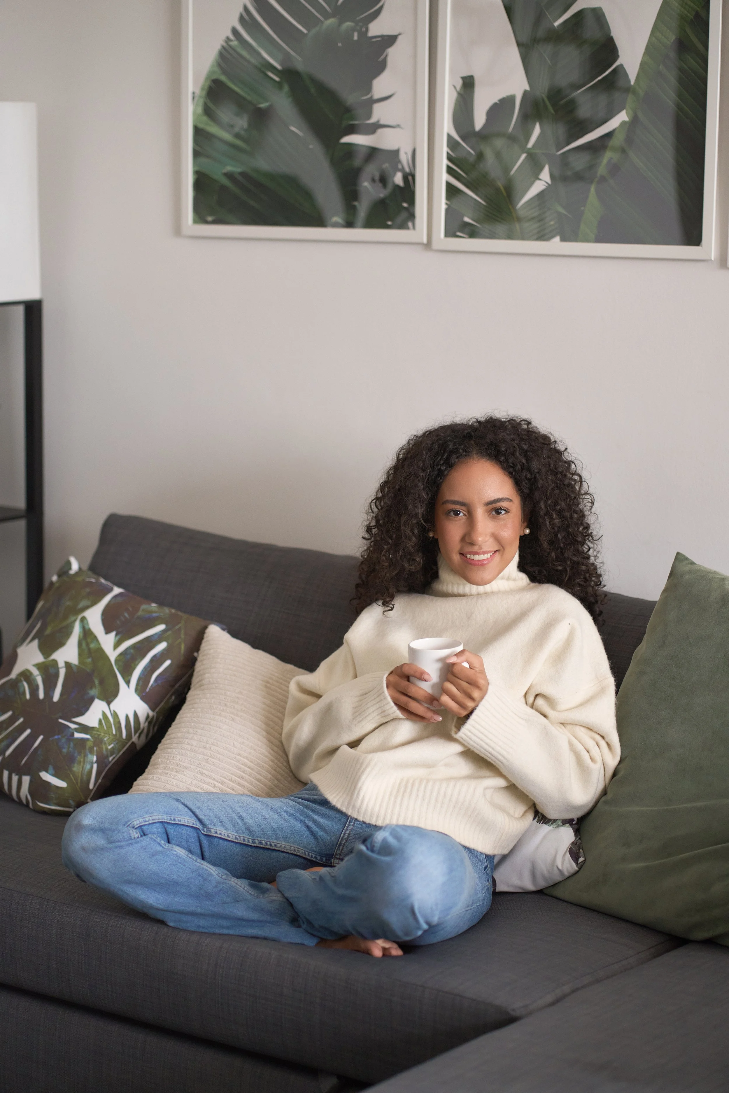 A woman with curly hair and light skin sitting on a gray couch in a living room, holding a white mug and smiling at the camera. She is wearing a cream-colored sweater and blue jeans. Behind her is a decorative pillow with a tropical leaf pattern. The background features a light gray wall with two framed botanical prints of green tropical leaves.