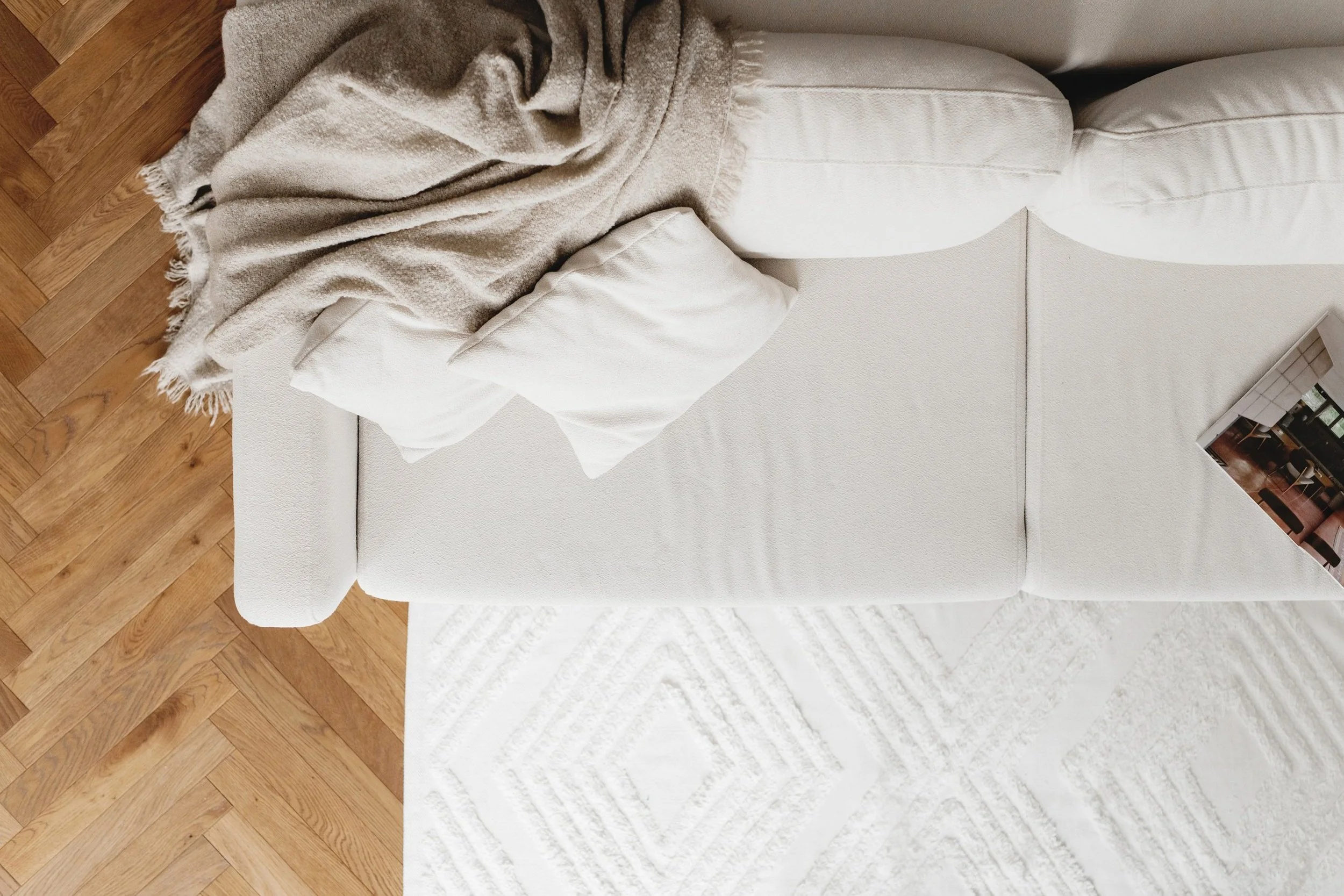 The image shows a beige sofa with a blanket draped over the armrest and a pillow, set on a hardwood floor. Part of a magazine or book is on the sofa's right side.