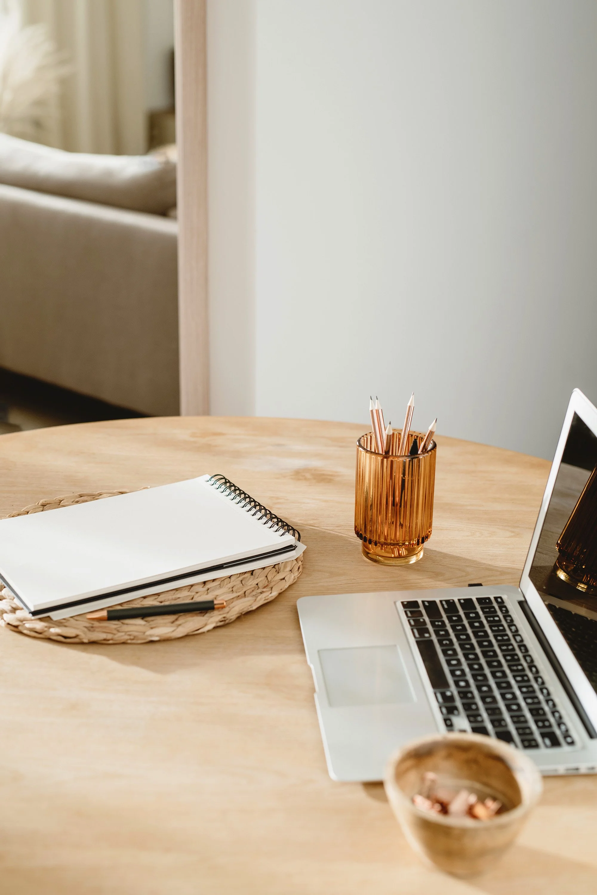 A wooden desk with a silver laptop, a spiral notebook on a woven basket, a black pen, a copper-colored pen holder with pens, a small bowl with nuts, and a glass of dark beverage. Part of a gray couch and a white wall are visible in the background.
