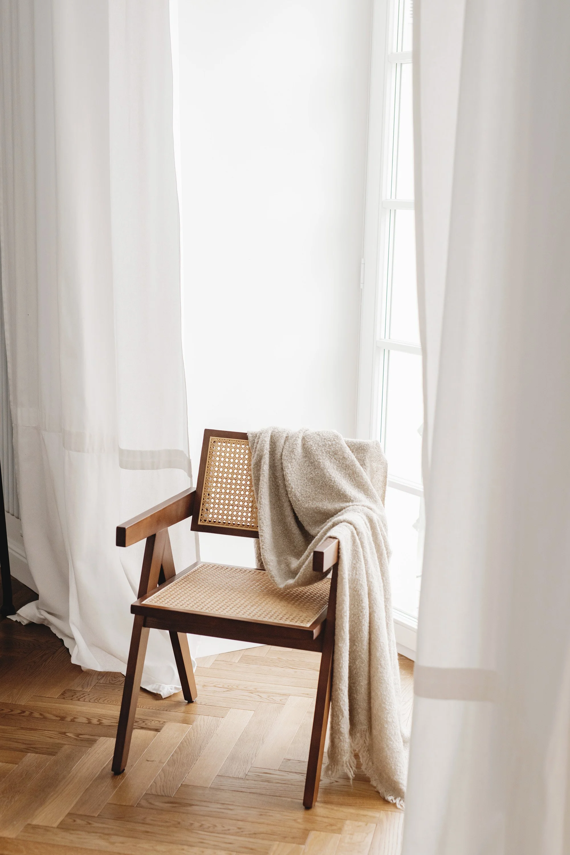 A wooden chair with a woven cane backrest and seat, draped with a beige blanket, in a bright room with light wood flooring, white curtains, and a large window allowing natural light.