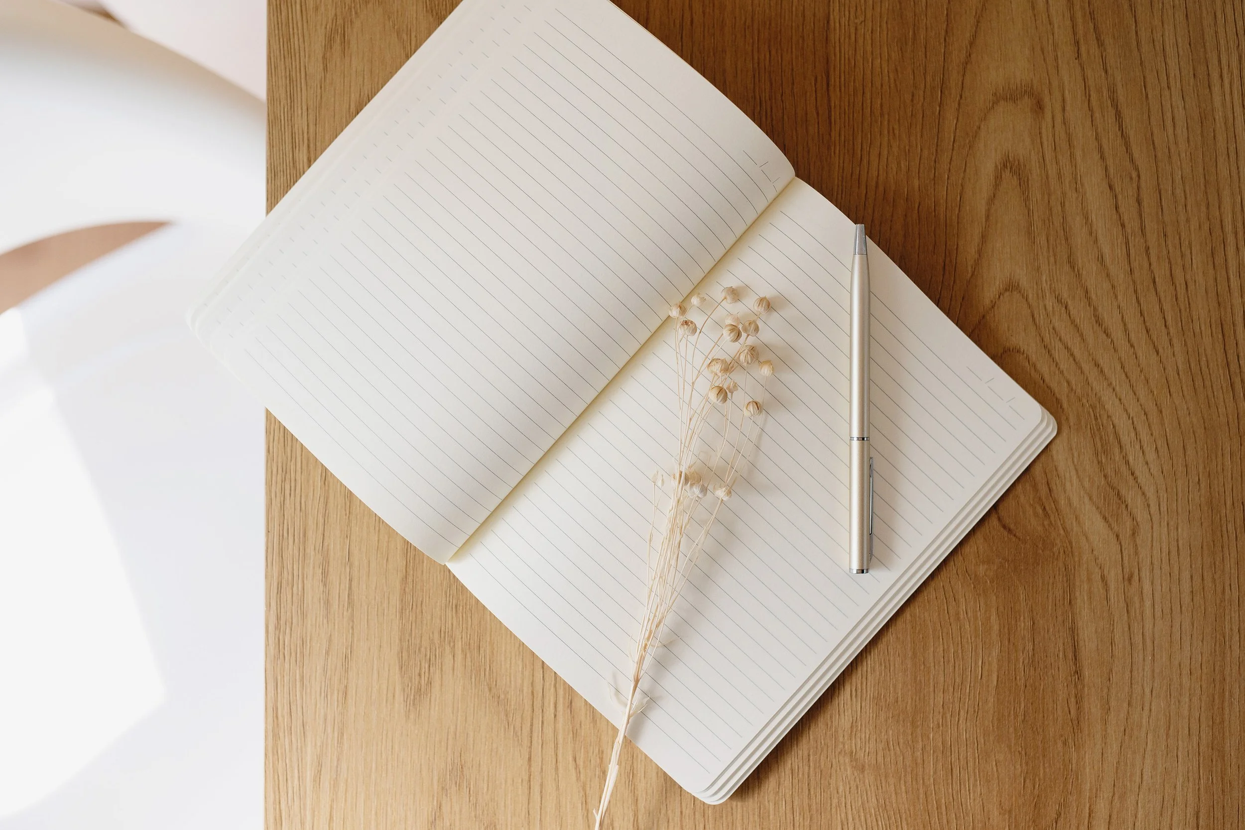 Open blank notebook with lined pages on wooden table, a silver pen beside it, and a small bunch of dried white flowers placed on the open pages.