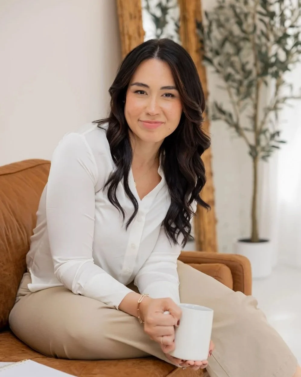 A woman sitting on a brown sofa holding a white mug, smiling, in a cozy room with a potted plant and curtains in the background.