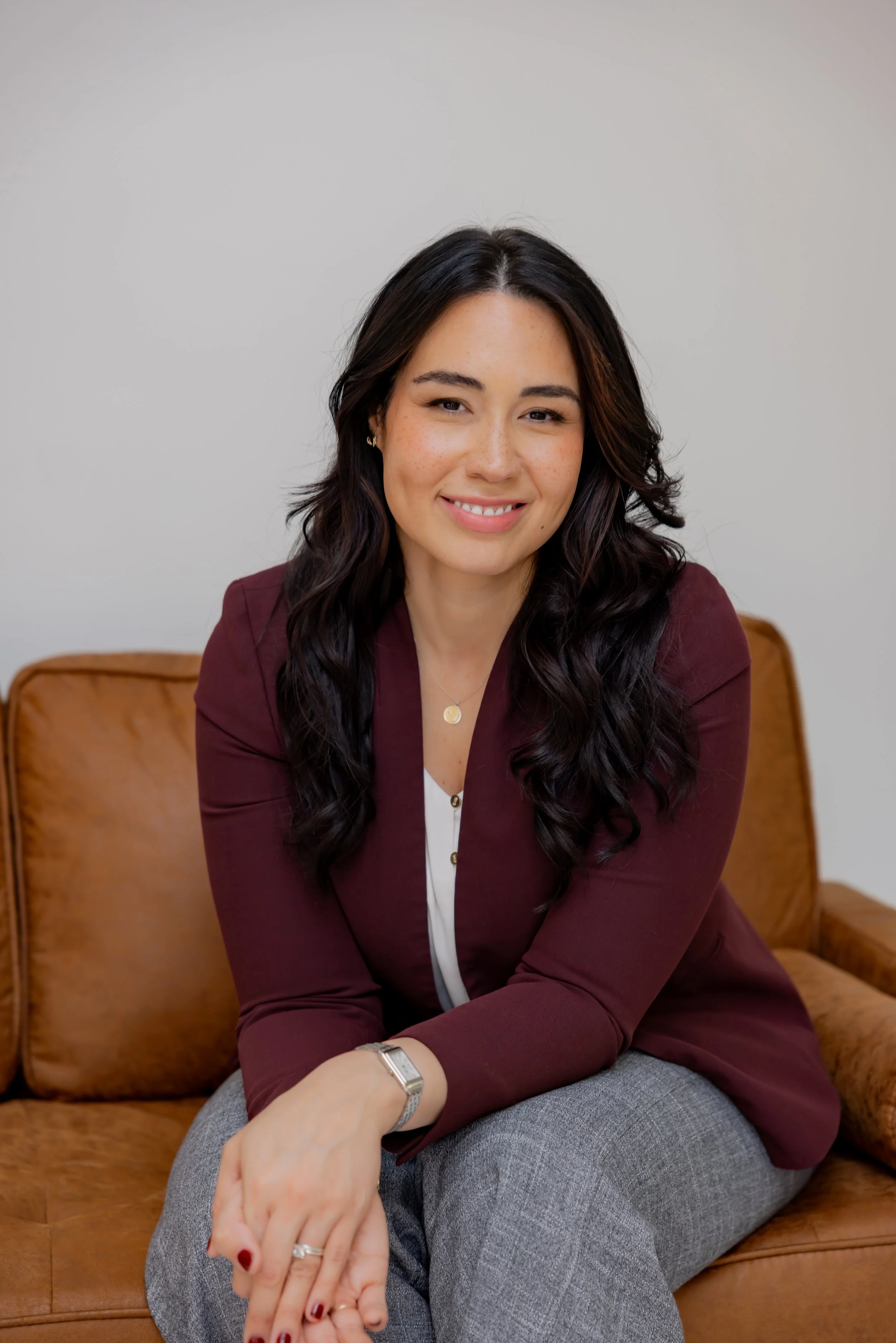 A woman sitting on a brown leather couch, smiling at the camera, wearing a maroon blazer, white shirt, gray pants, and jewelry, with dark wavy hair and a neutral background.