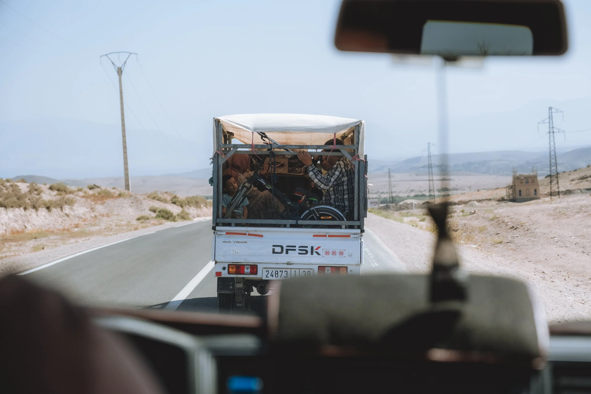 Une vue depuis l'intérieur d'un véhicule montrant une route désertique et un camion en détresse avec des personnes à l'arrière, dans un paysage aride avec des pylônes électriques et un bâtiment en arrière-plan.